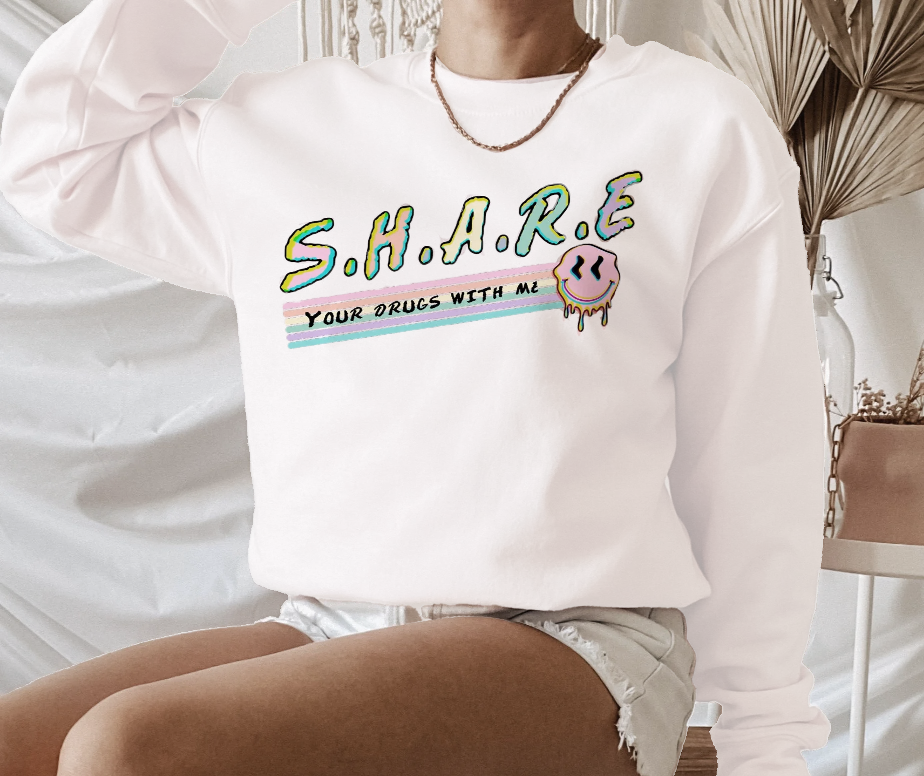 A person sits on a chair in a HiCiti S.H.A.R.E Your Drugs With Me Sweatshirt, a unisex crewneck with colorful text and a melting smiley face. Neutral decor and dried plants in the background create a relaxed atmosphere.