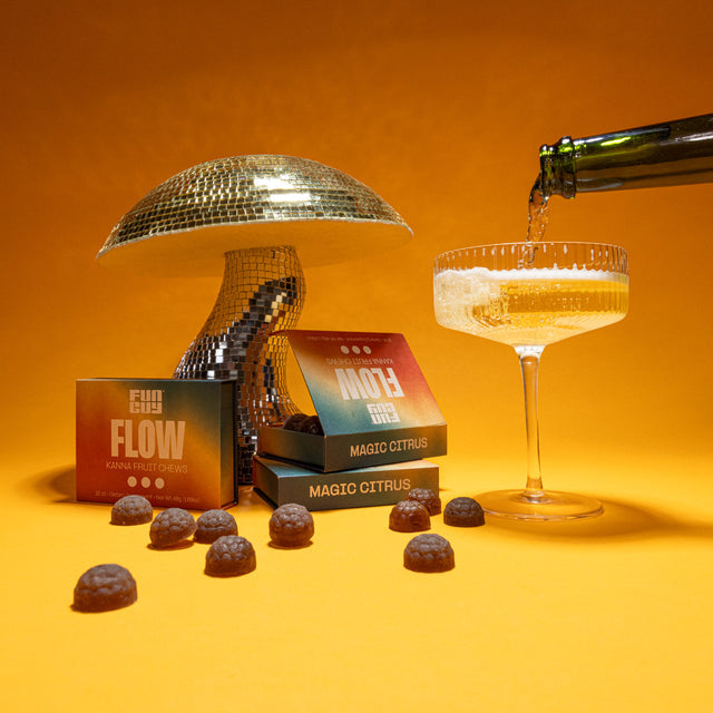 A hand pours sparkling liquid into a coupe glass next to a mushroom disco ball, Fun Guy Flow Fruit Chews boxes, and colorful chews—all set against a vibrant orange backdrop, celebrating these natural mood-enhancing treats.