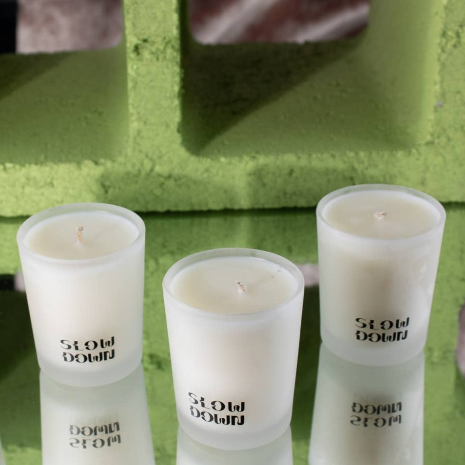 The CANDLE VOTIVE SET 3 - COCONUT WAX by Slow Down features three white coconut wax candles in frosted glass holders, arranged on a reflective surface before a green cinder block for a tranquil touch.