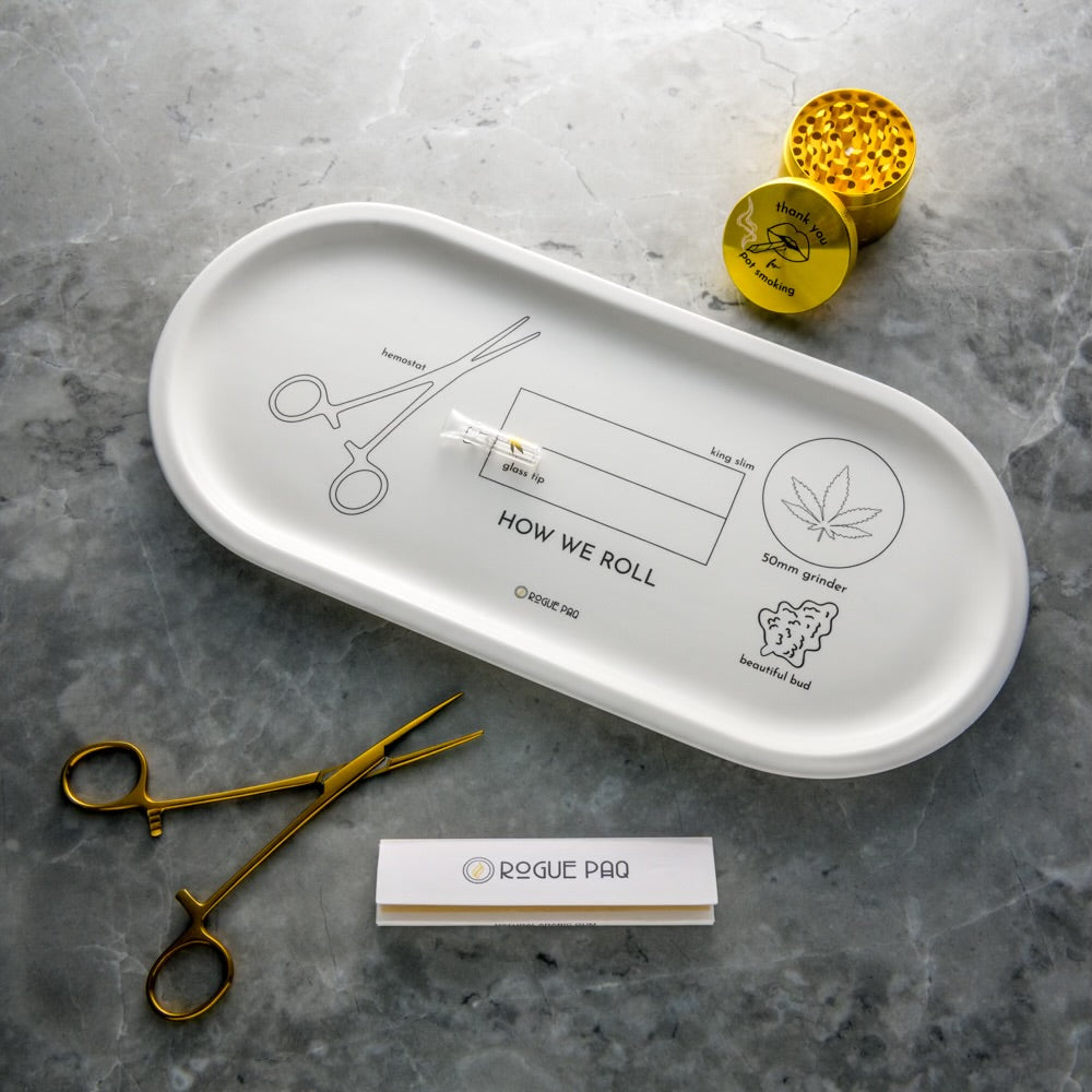 The How We Roll Porcelain Valet Tray by Rogue Paq displays rolling tool diagrams alongside gold scissors, a gold grinder, and a branded rolling paper pack—all arranged on gray marble for a stylish entryway accent.