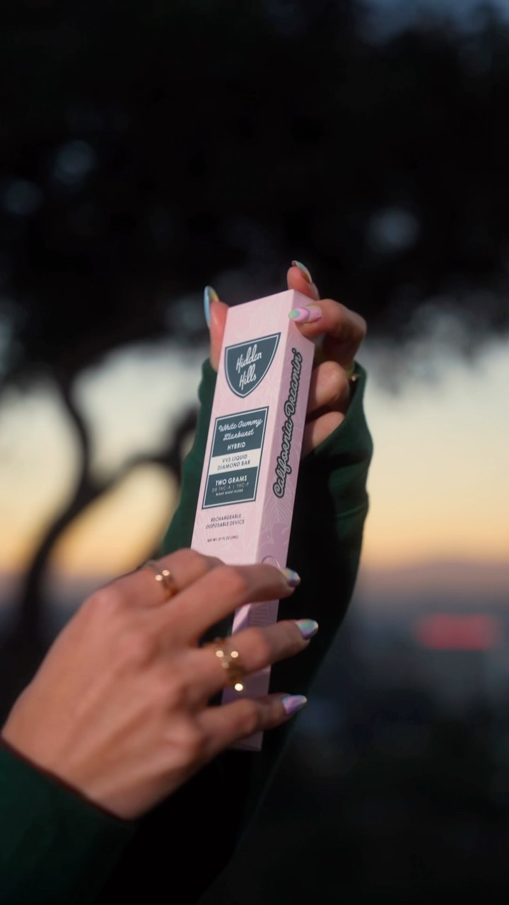 A person with manicured nails and gold rings holds a pink and green Hidden Hills Liquid Diamonds Disposable box outdoors, with trees and a sunset blurred in the background.