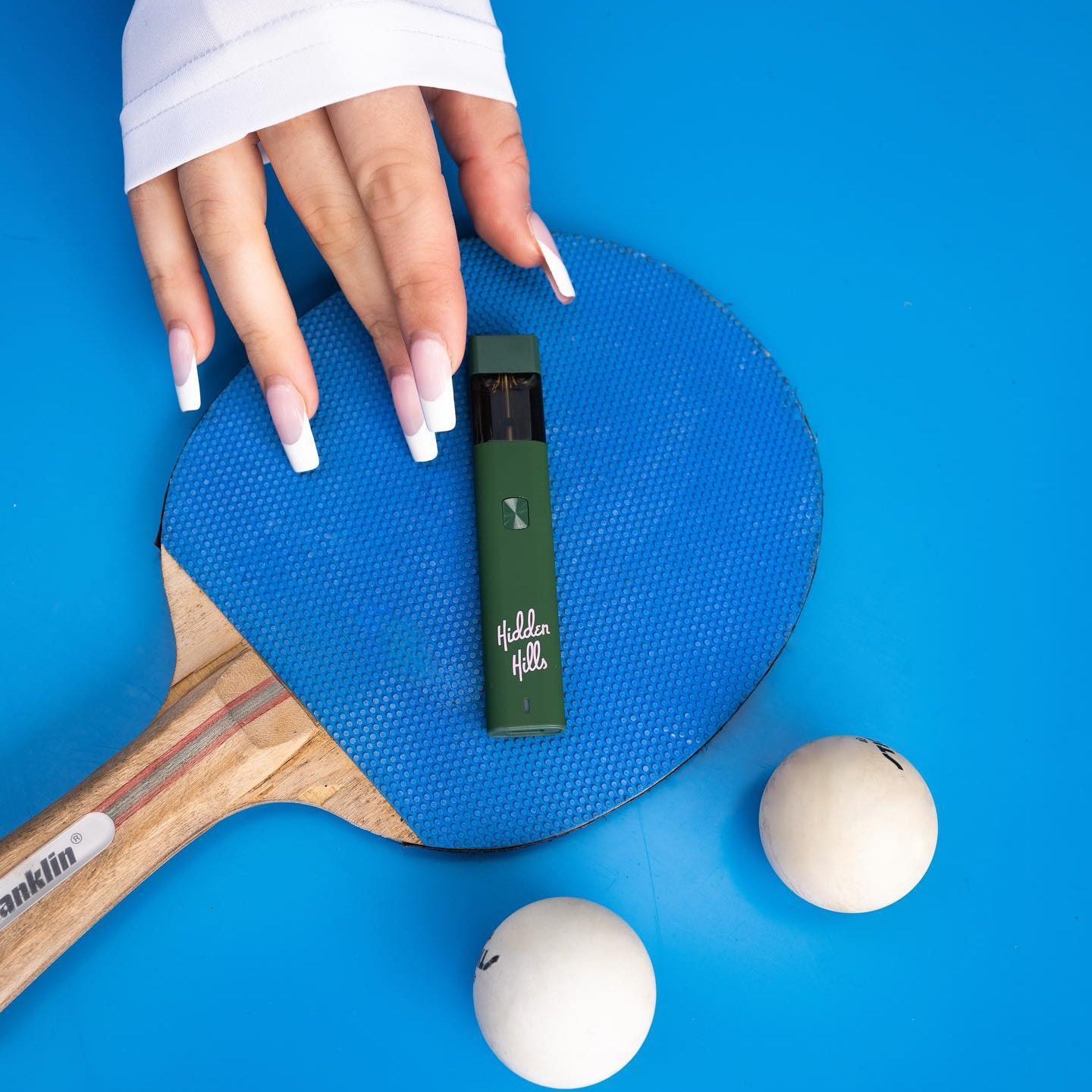 A hand with long, white acrylic nails touches a blue ping pong paddle topped with a Hidden Hills Liquid Diamonds Disposable vape pen. Two white ping pong balls sit nearby on the bright blue surface.