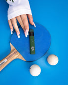 A hand with long, white acrylic nails touches a blue ping pong paddle topped with a Hidden Hills Liquid Diamonds Disposable vape pen. Two white ping pong balls sit nearby on the bright blue surface.