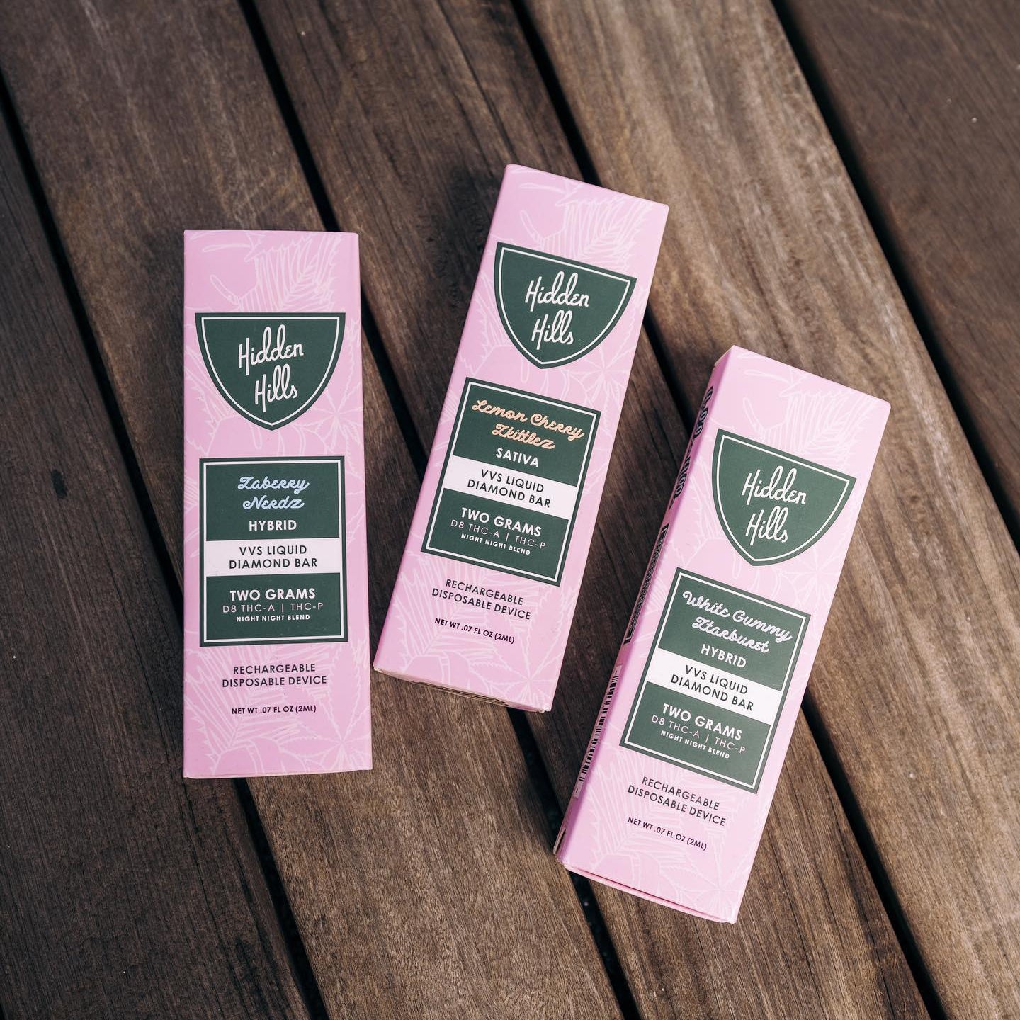 Three Hidden Hills Liquid Diamonds Disposable vape pen boxes in pink and green are displayed on wood. Each box highlights unique cannabis strains, Delta 8 content, and bold details.