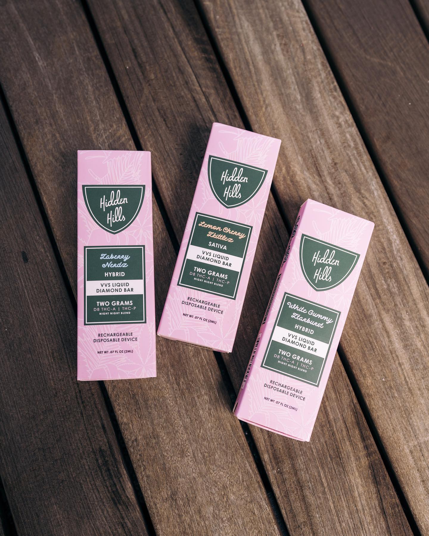 Three Hidden Hills Liquid Diamonds Disposable vape pen boxes in pink and green are displayed on wood. Each box highlights unique cannabis strains, Delta 8 content, and bold details.