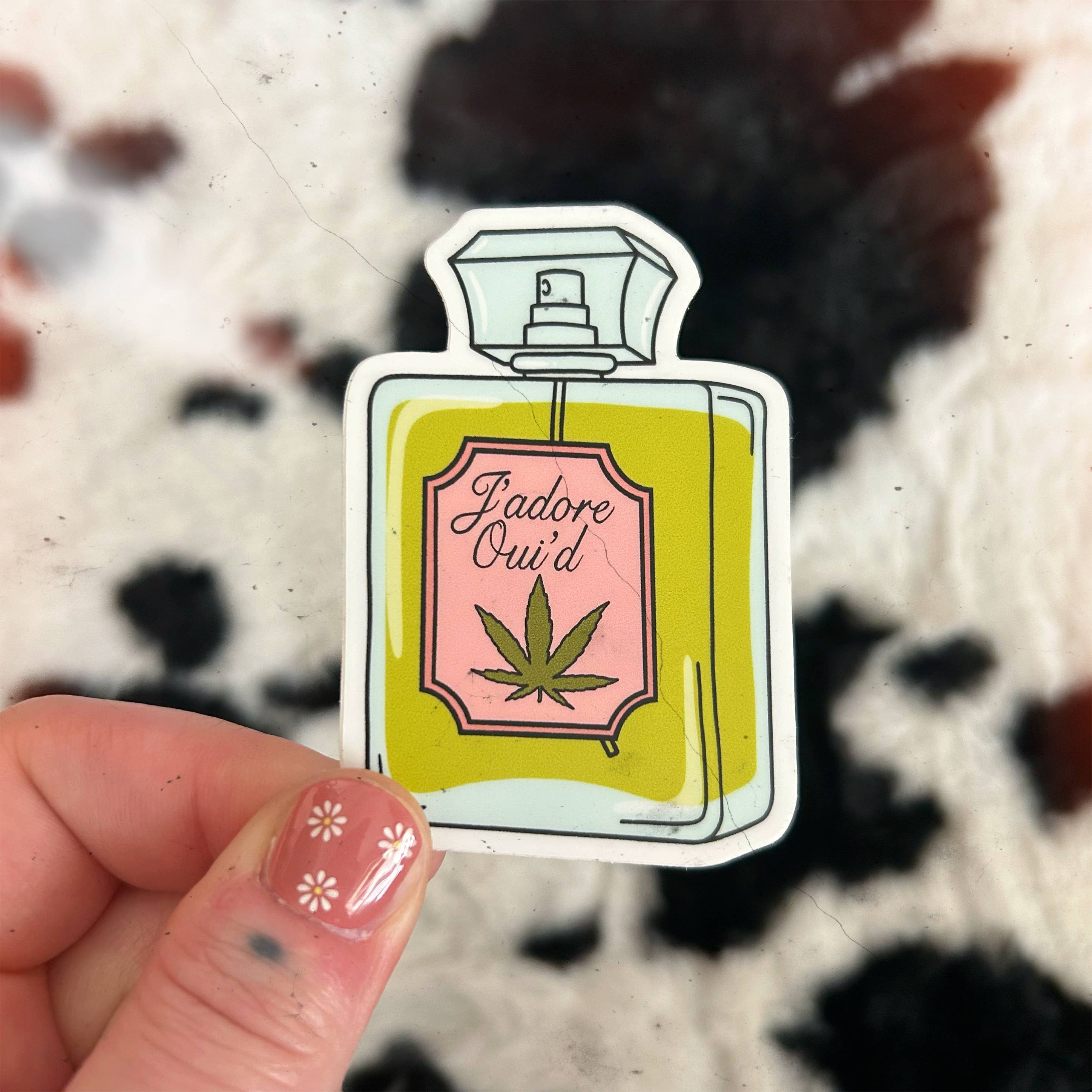 A hand with floral nail art holds the Jadore Oui’d Sticker by Chateau Blanche—a matte perfume bottle design with a cannabis leaf. The fuzzy, black-and-white spotted background adds charm to this playful sticker moment.