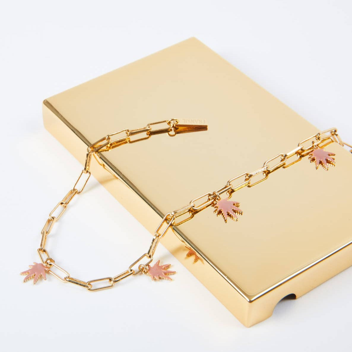 The Frankie Smoke Leaf Charm Necklace, featuring pink and gold canna-leaf charms, is displayed draped over a rectangular gold box against a white background.