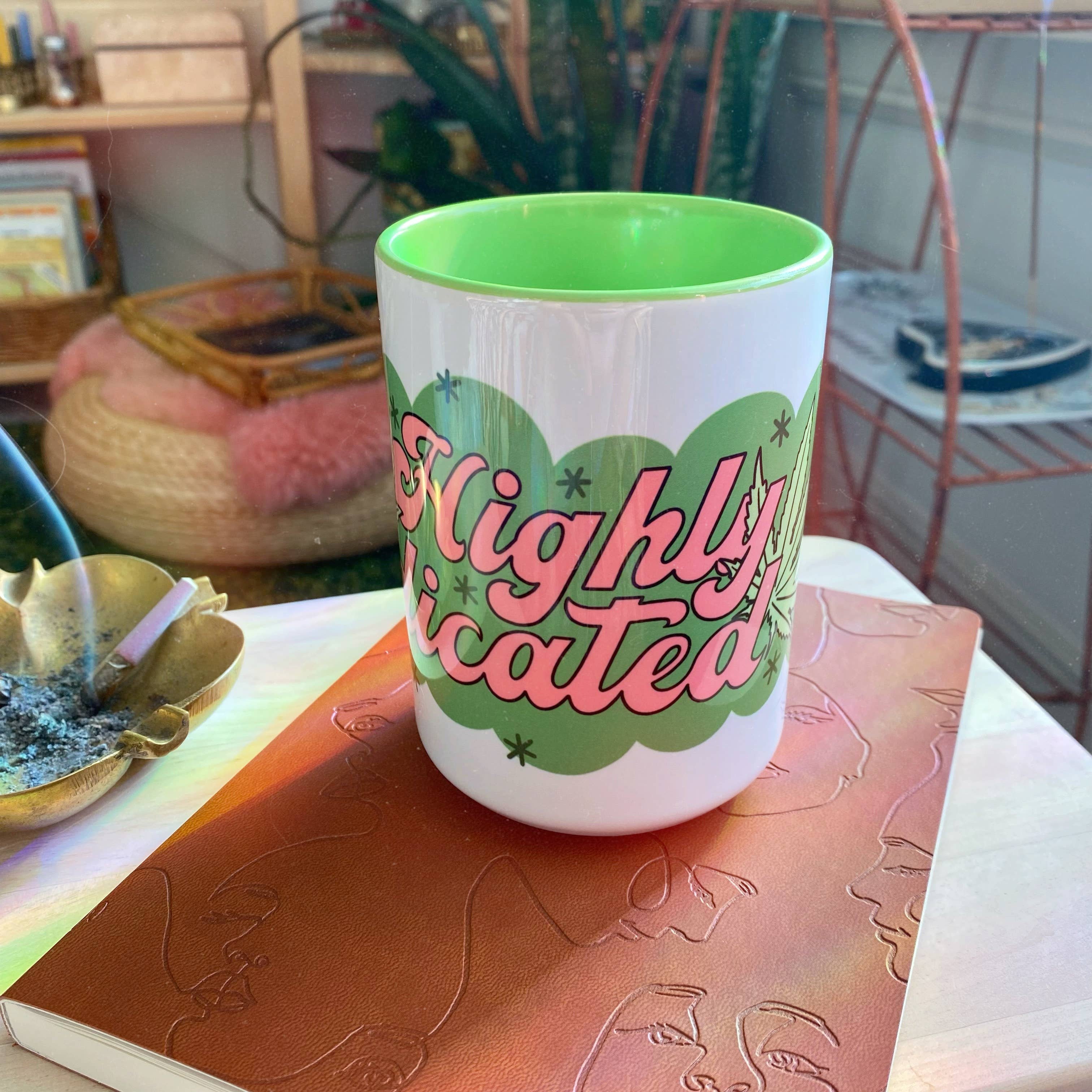 The Chateau Blanche Coffee Mug Highly Medicated features pink and green text on white with a green interior. Shown atop a brown embossed notebook, its surrounded by plants, books, and a burning incense stick for a cozy, 420-friendly vibe.