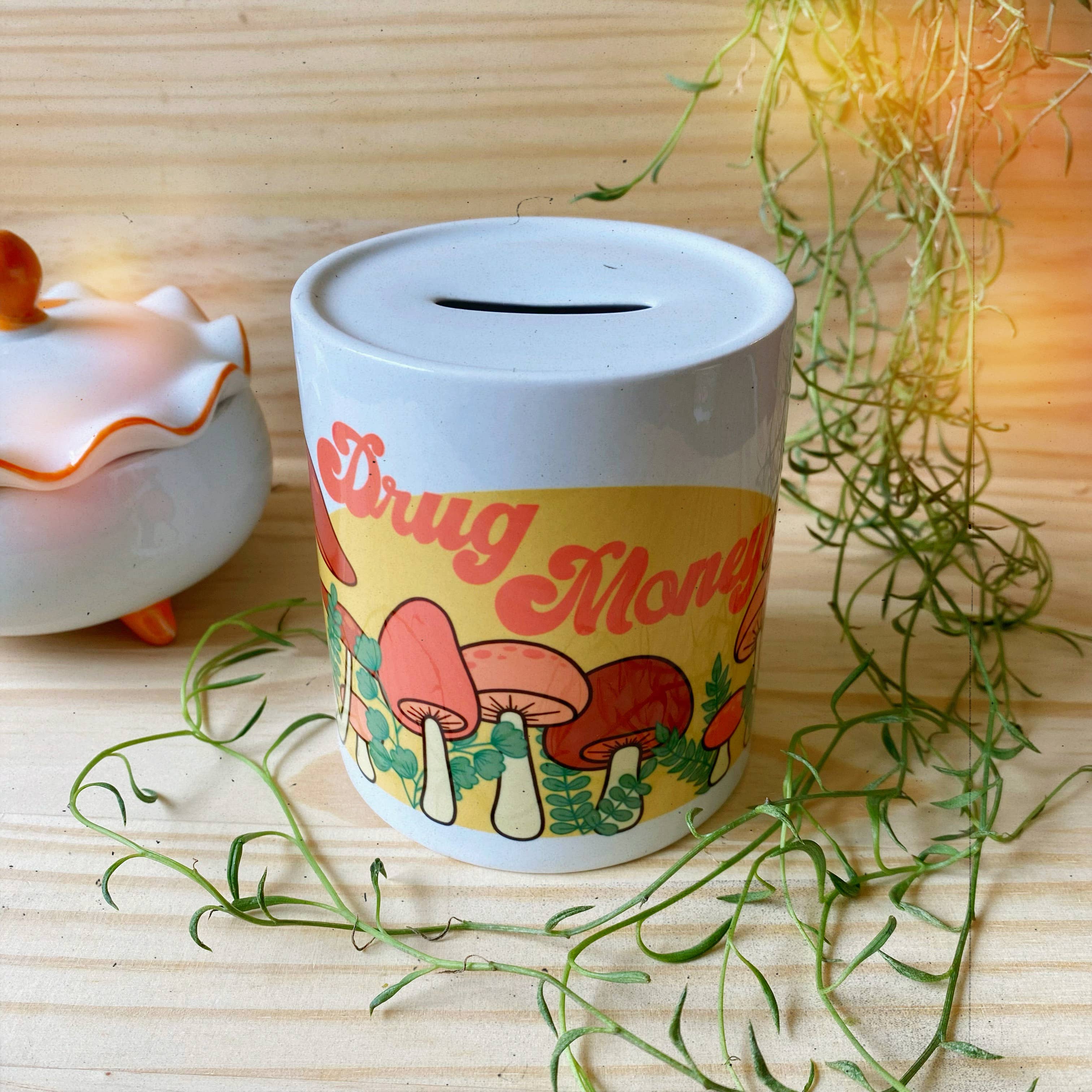 The Chateau Blanche Drug Money Piggy Bank with Mushrooms, featuring psychedelic mushroom illustrations and Drug Money text, sits on a wooden surface near a white container with an orange lid, surrounded by green plants.