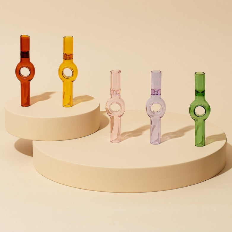 Four vibrant Charlotte Pipes by Laundry Day—orange, pink, purple, and green—stand upright on two cream round platforms against a beige background. Each glass pipe features a circular center detail.