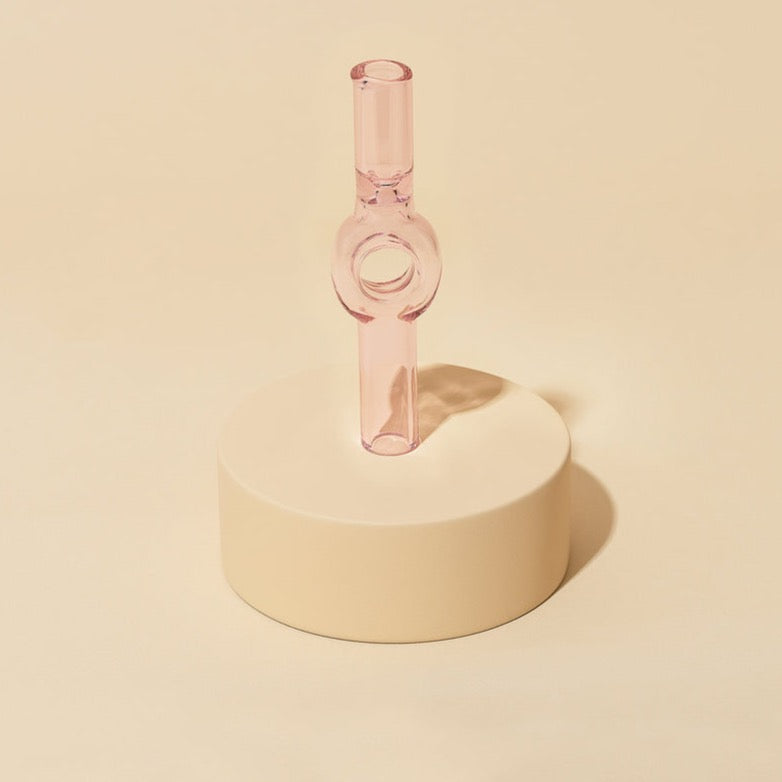 The Charlotte Pipe by Laundry Day is a pink, translucent, hand-blown tube-shaped pipe with a round opening, standing upright on a smooth beige pedestal against a matching background.