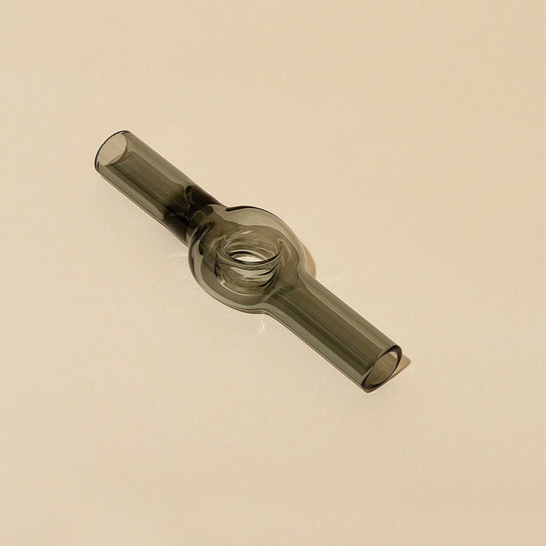 The Laundry Day Charlotte Pipe is a transparent, cylindrical glass one-hitter with a bulbous center, open ends, and a smooth modern design. It is shown on a plain beige background, highlighting its symmetrical appearance.