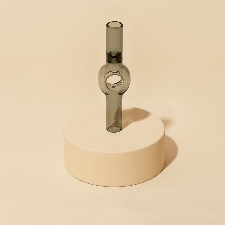The Charlotte Pipe by Laundry Day is a minimalist, gray hand-blown pipe with a circular center and two vertical ends, displayed on a smooth beige platform against a matching background.