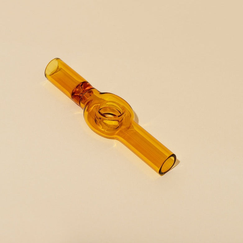 The Charlotte Pipe by Laundry Day is a translucent amber hand-blown piece with a circular center and straight tubes on each side, shown on a light beige surface.