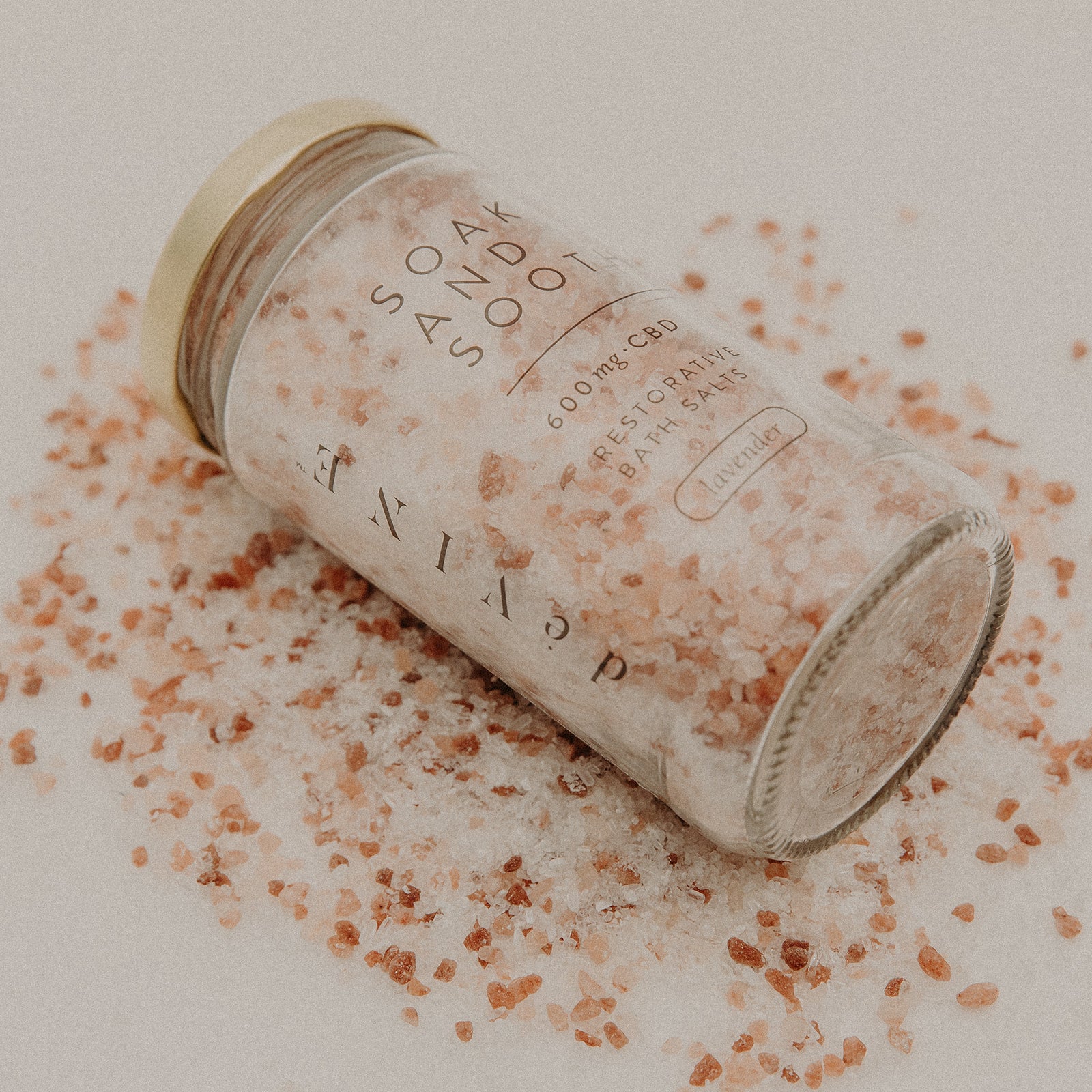 A jar of DeVINE Wellness Soak & Soothe Bath Salts with epsom salt and pink Himalayan crystals spills onto a light surface, creating an inviting atmosphere of relaxation.