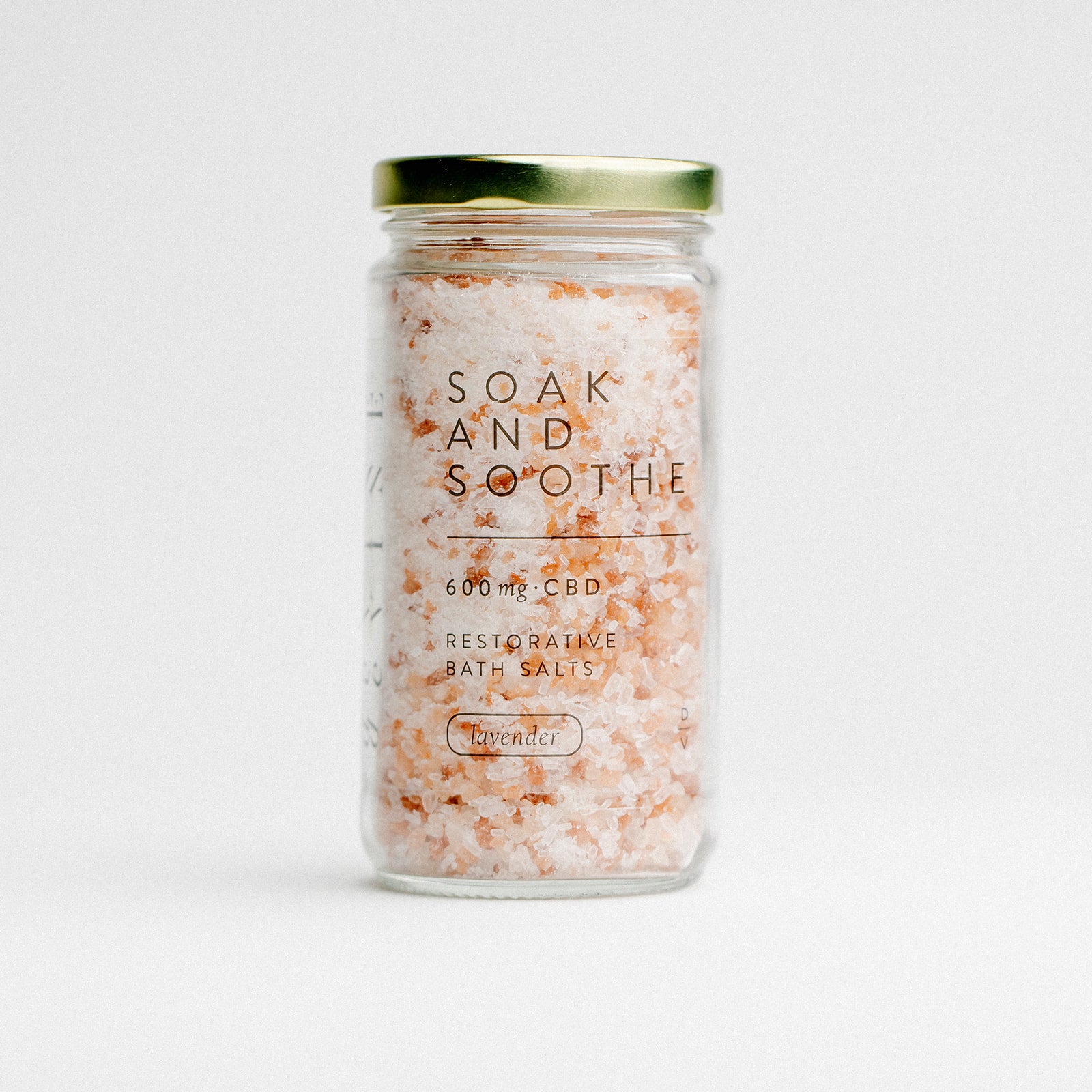 A clear glass jar with a gold lid contains pink and white Soak & Soothe Bath Salts by DeVINE Wellness—600 mg CBD, made with epsom salt and lavender. The label says Restorative Bath Salts. Photographed against a white background.