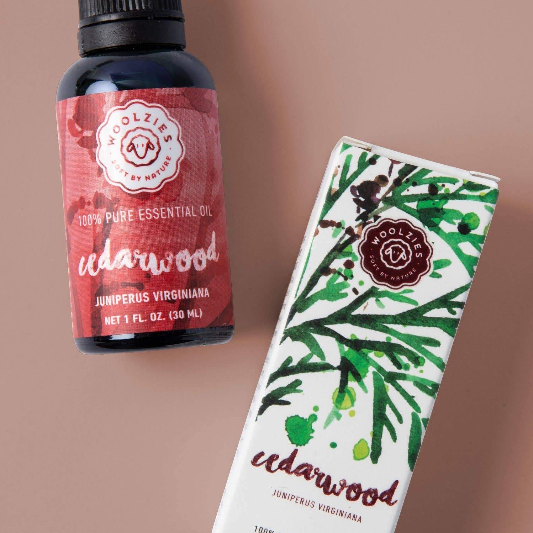 A bottle of Woolzies Cedarwood Essential Oil, a soothing and natural insect repellent, sits beside its box decorated with green cedar branch designs. The red label contrasts with the white boxs elegant green artwork.