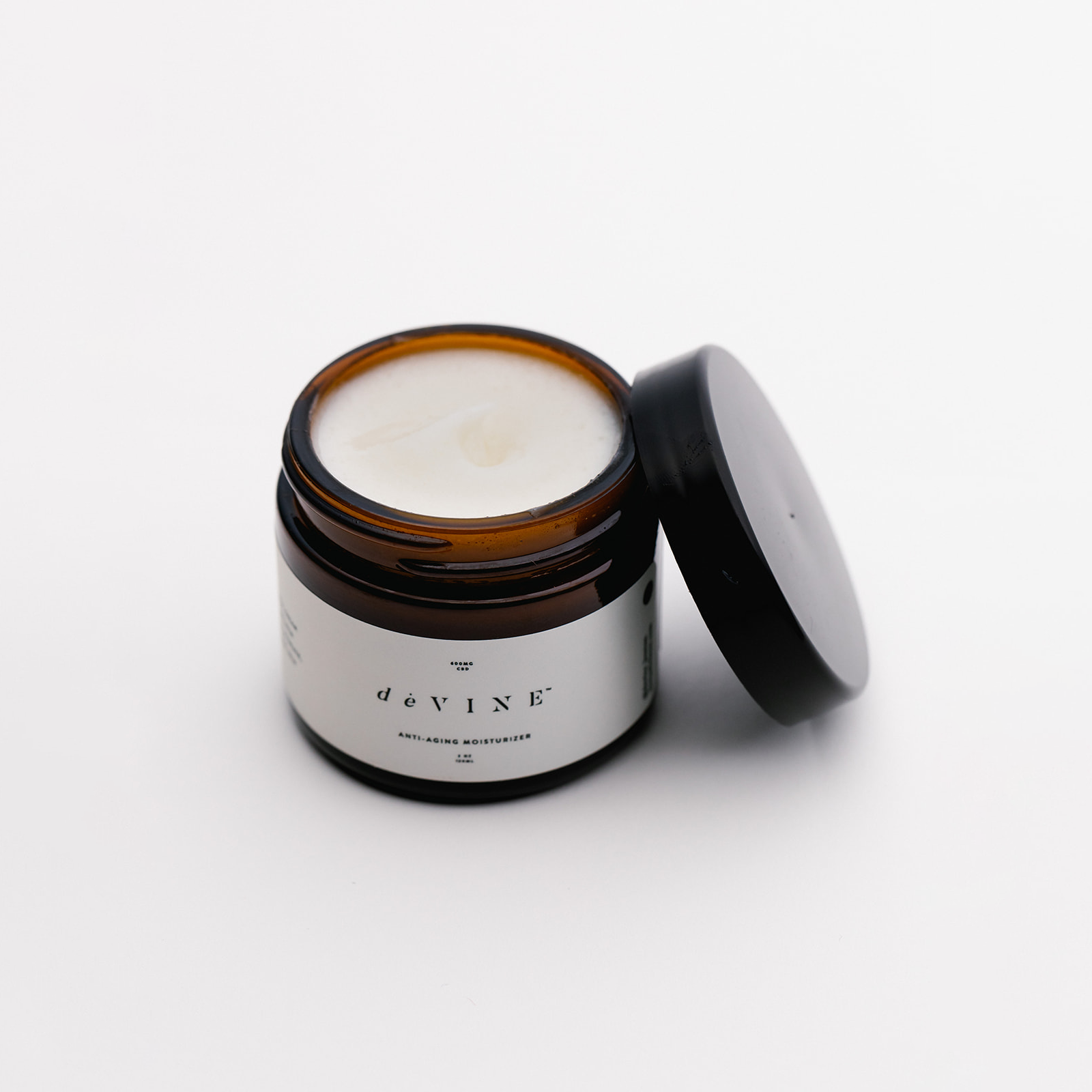 A brown glass jar of DeVINE Wellness deVINE Anti Aging Moisturizer sits open on a white surface, its black lid nearby. The smooth, white cream inside offers rich hydration and anti-aging benefits for your skin.