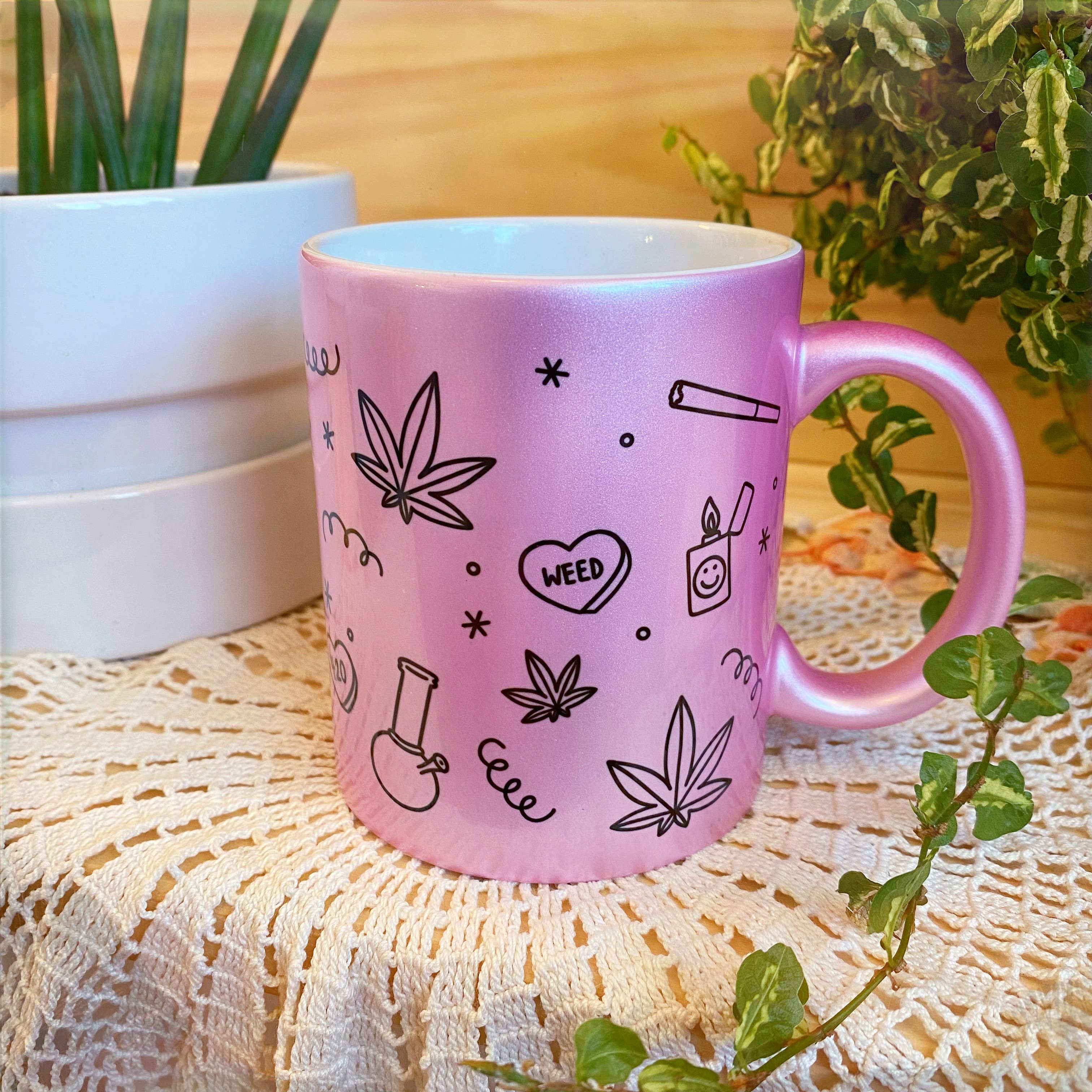 A handmade Chateau Blanche Metallic Paraphernalia Coffee Mug, pink with black doodles of marijuana leaves, a bong, a joint, and the word WEED, sits on a crochet doily near green plants—perfect for your quirky mug collection.