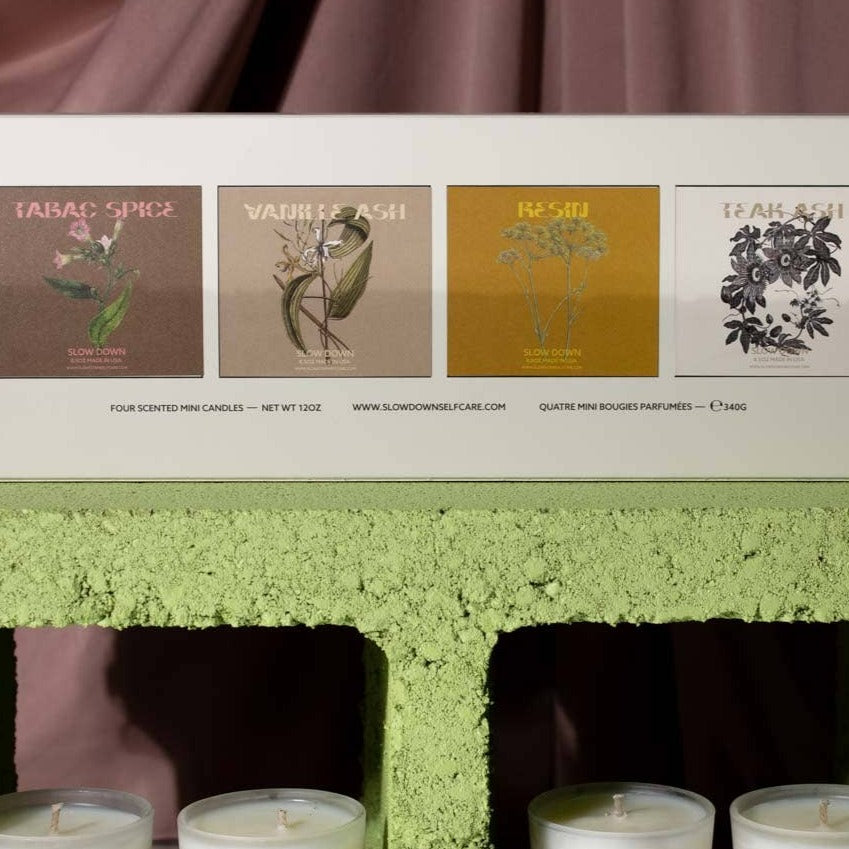 A white box with four illustrated labels—Tabac Spice, Vanilla Cash, Resin, and Teak Sili—rests on a green block, showcasing Slow Downs CANDLE VOTIVE SET 3 - COCONUT WAX candles partially visible below.
