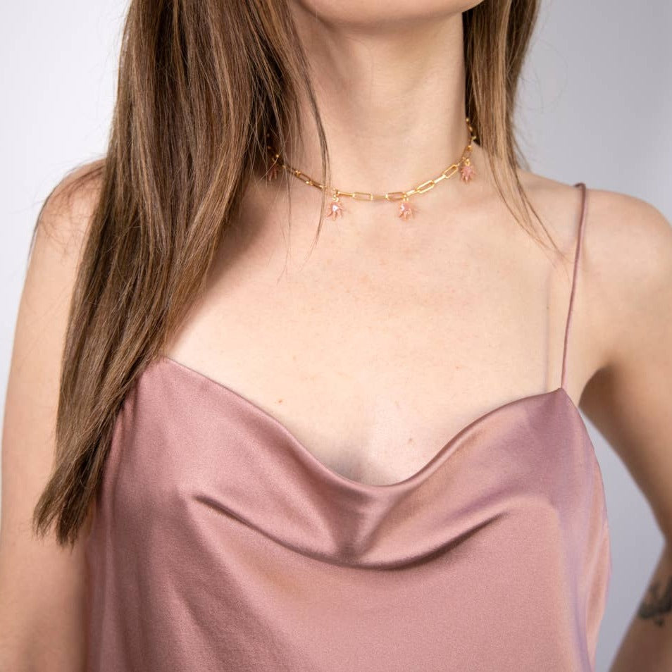 A woman wears a mauve satin spaghetti strap top and the Frankie Smoke Leaf Charm Necklace—an adjustable gold paperclip chain with small pink charms—shown from shoulders to just above her lips.