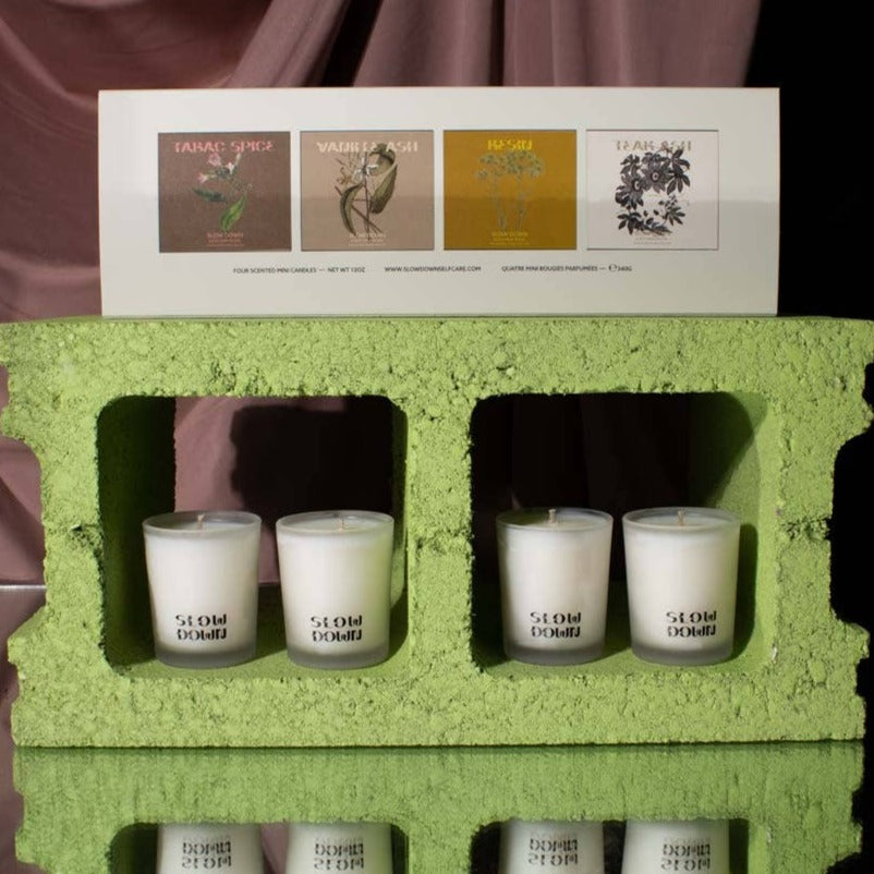Four Slow Down CANDLE VOTIVE SET 3 - COCONUT WAX votives in glass holders rest in a green cinder block. A card shows the sets fragrance names and illustrations: Farag Spice, Vanillaash, Roseau, and Tisane.