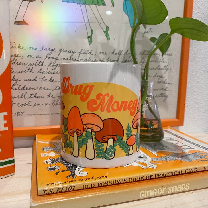 The Chateau Blanche Drug Money Piggy Bank with Mushrooms features orange and yellow text on a white mug-style bank, decorated with psychedelic mushrooms and shown among books and botanical decor.
