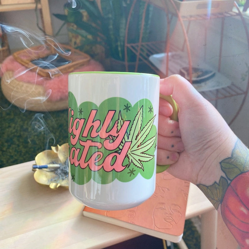 A hand holds the Chateau Blanche Coffee Mug Highly Medicated, featuring a cannabis leaf. Smoke rises from incense on a table with plants and cozy decor, creating a chill, 420-friendly atmosphere.
