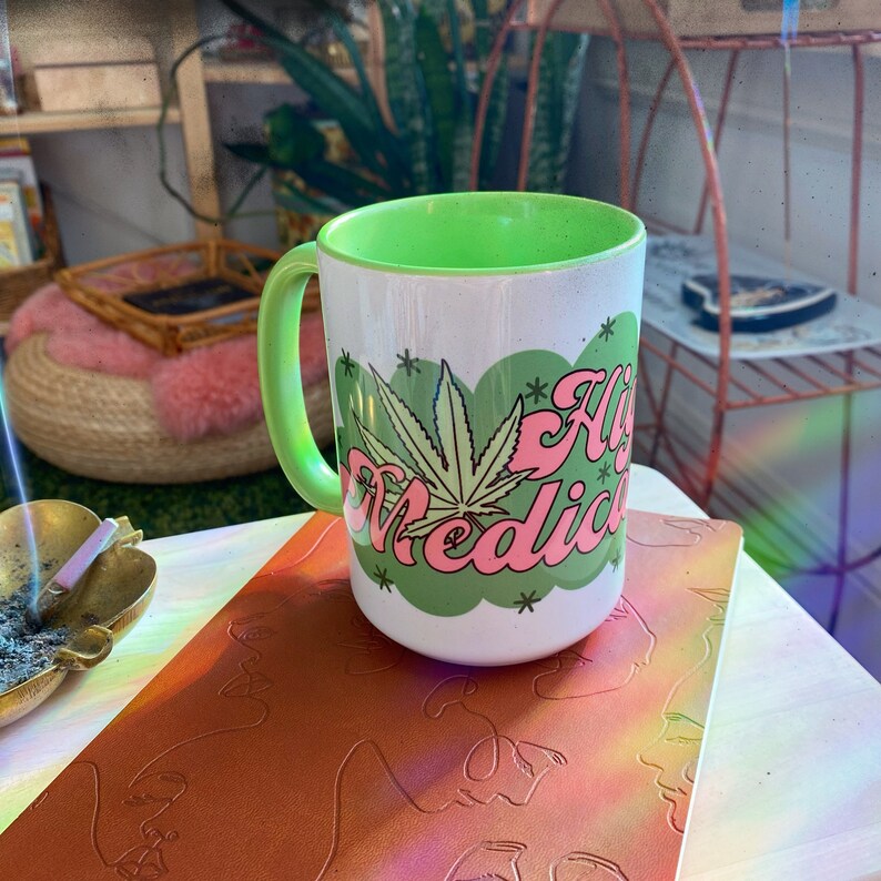 A white and green Chateau Blanche Coffee Mug Highly Medicated with a cannabis leaf sits on a metallic embossed notebook, with an ashtray nearby and a cozy, 420-friendly room featuring plants and shelves in the background.