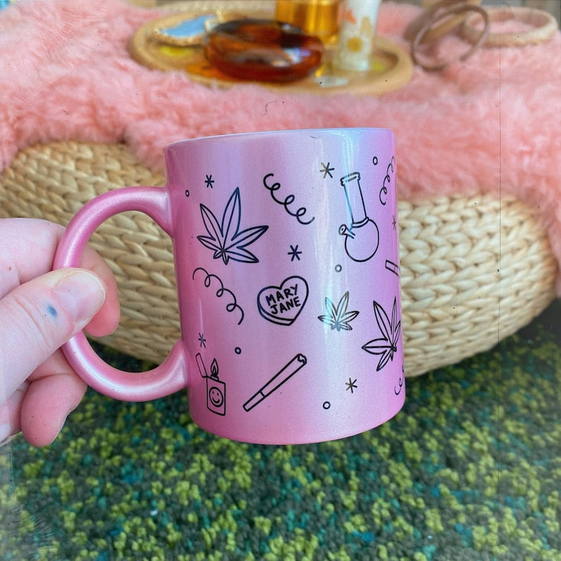 A hand holds the Chateau Blanche Metallic Paraphernalia Coffee Mug, featuring marijuana leaves, a bong, lighter, vape pen, swirls, and a Mary Jane heart. Handmade in Tulsa, it sits on a woven table with fuzzy pink fabric behind.
