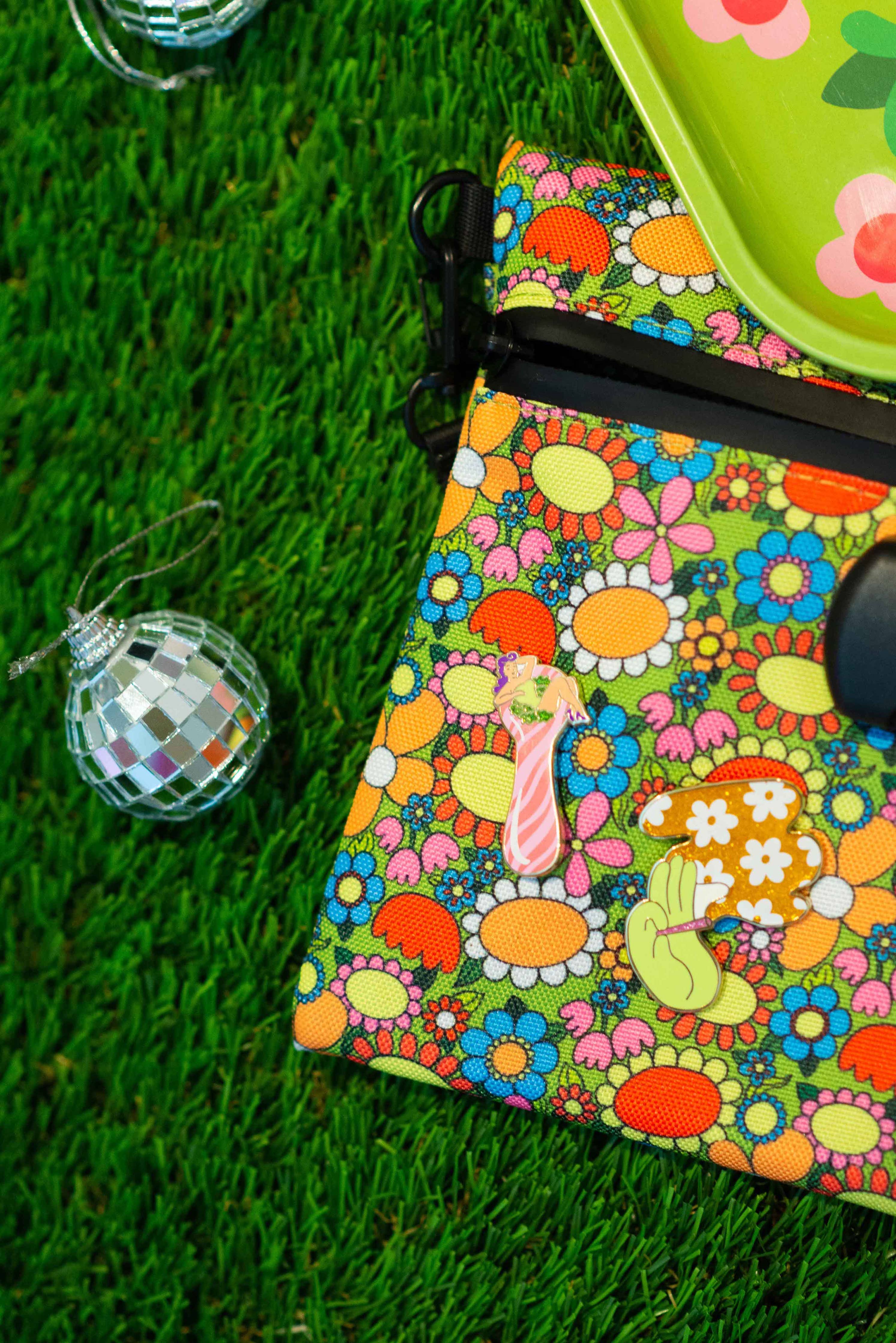 A small pouch with a colorful floral pattern and quirky pins, including The Peach Fuzz’s Total Smoke Show Enamel Pin, rests on green grass beside a shiny disco ball ornament and a flower-designed green tray.