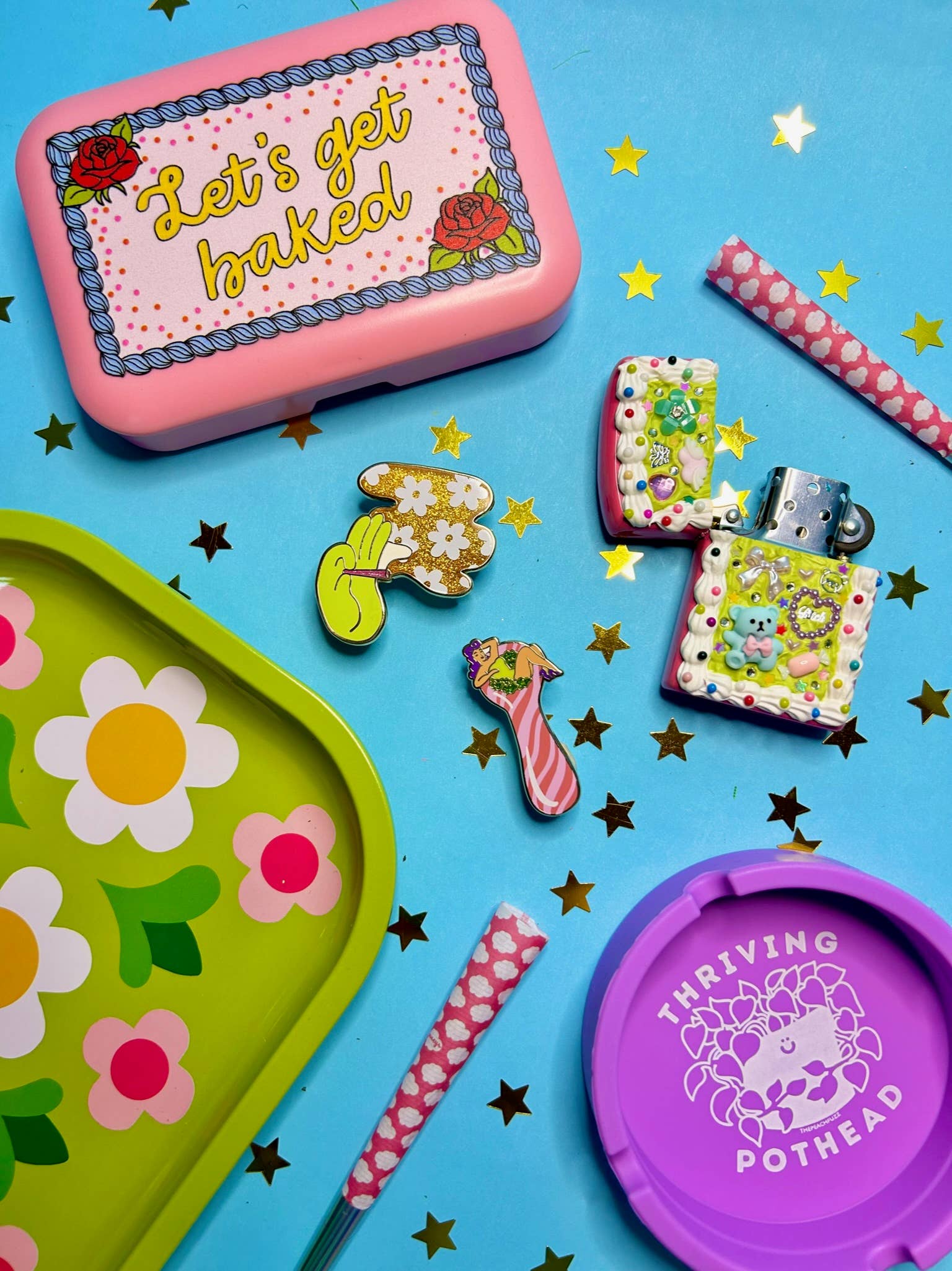 Colorful flat lay with a pink Lets Get Baked tin, green floral tray, purple Thriving Pothead container, decorated lighters, The Peach Fuzz Total Smoke Show Enamel Pin, star confetti, glitter pin accents, and pink polka-dot straws on a blue background.