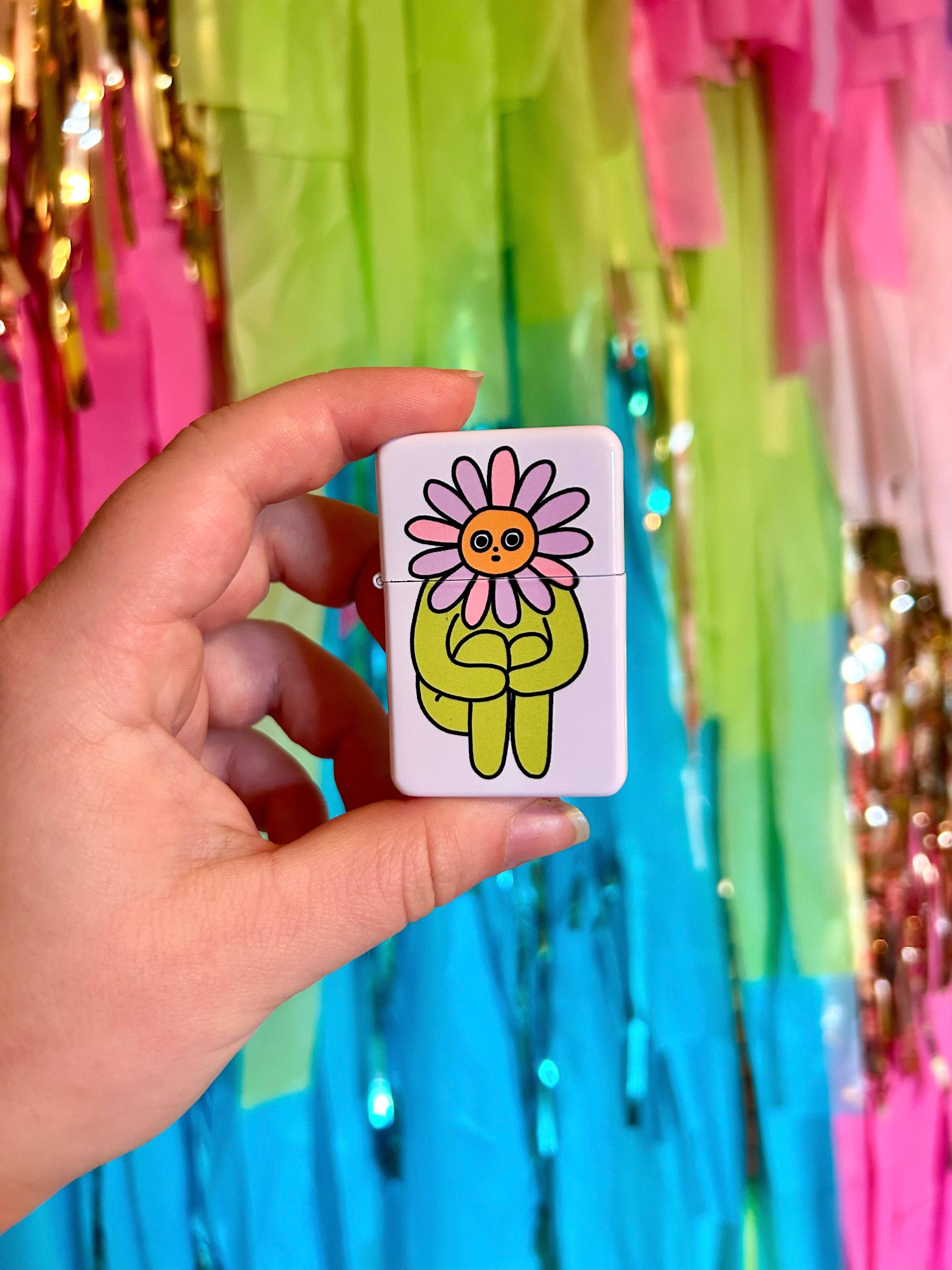 A hand holds The Peach Fuzz’s Flower Boy Lighter: Perfect, a refillable lighter with a cartoon pink daisy face and green body. Colorful fringed streamers burst in the background, making it a fun gift for stoners or candle lovers.