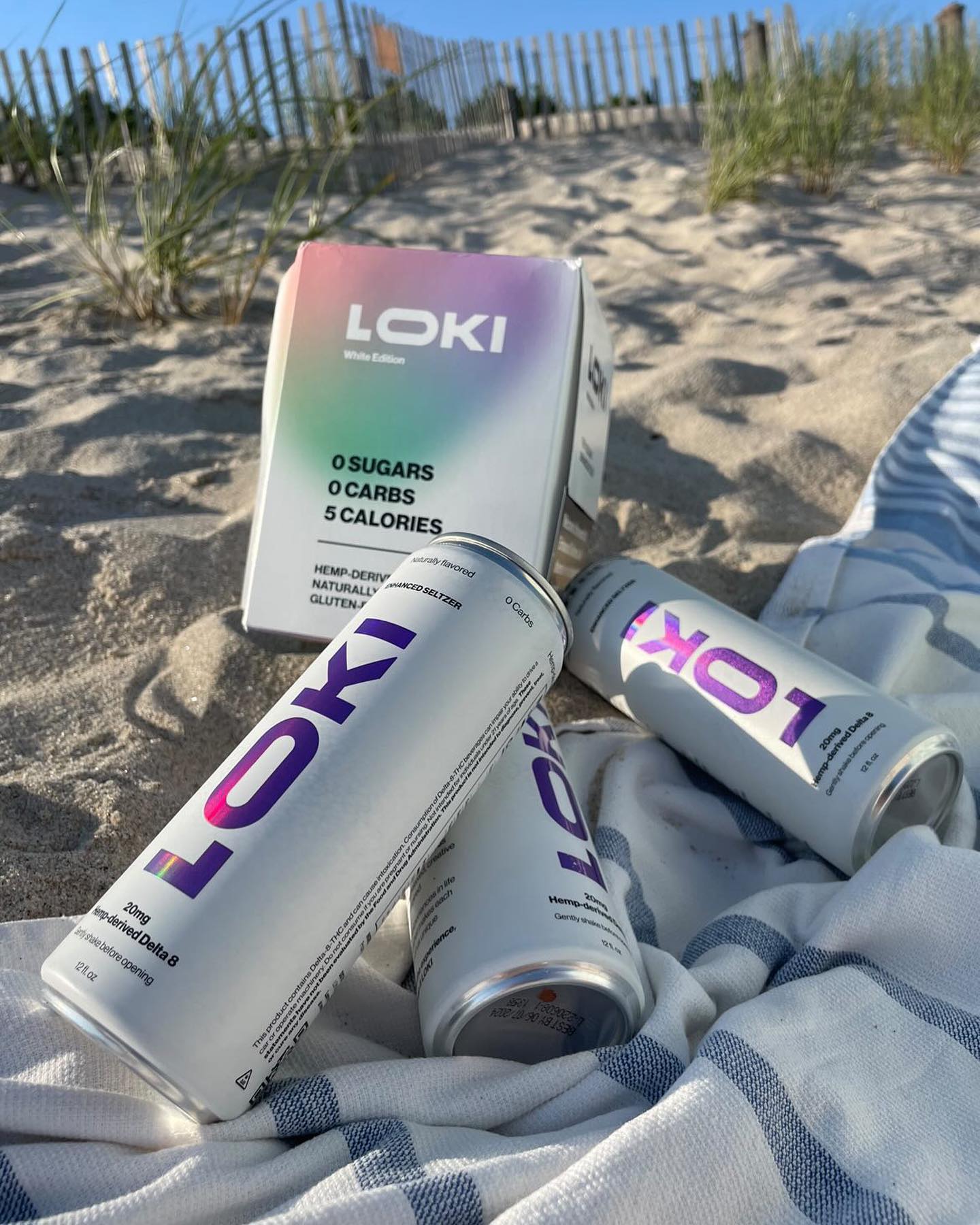 Three LOKI Seltzer cans and a LOKI box sit on a sandy beach near a striped towel, with grass and a wooden fence in the background under a bright, sunny sky.