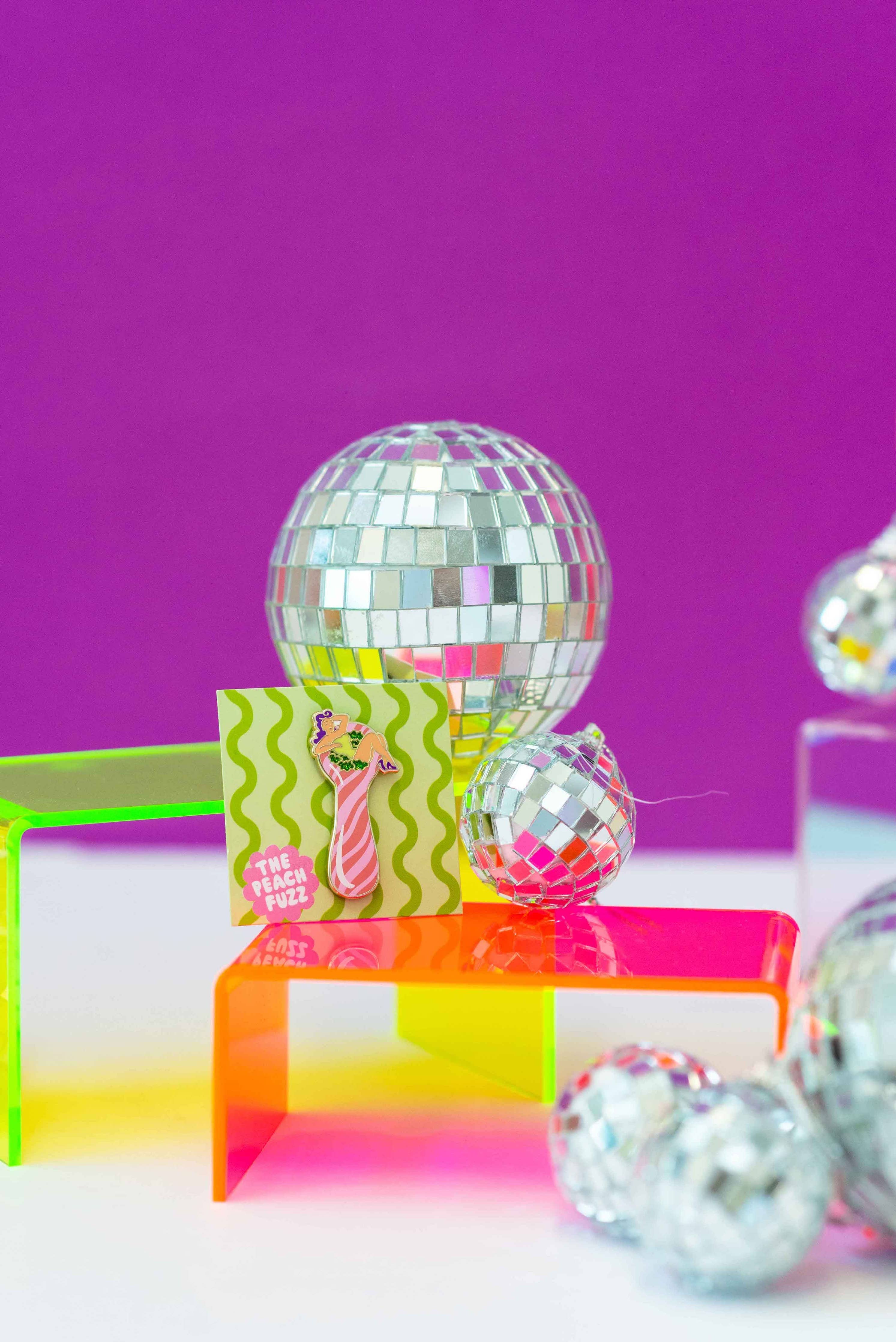 A collection of reflective disco balls in various sizes sits on neon orange and green acrylic stands, with The Peach Fuzzs Total Smoke Show Enamel Pin and a small colorful card in front, all set against a vibrant purple background.