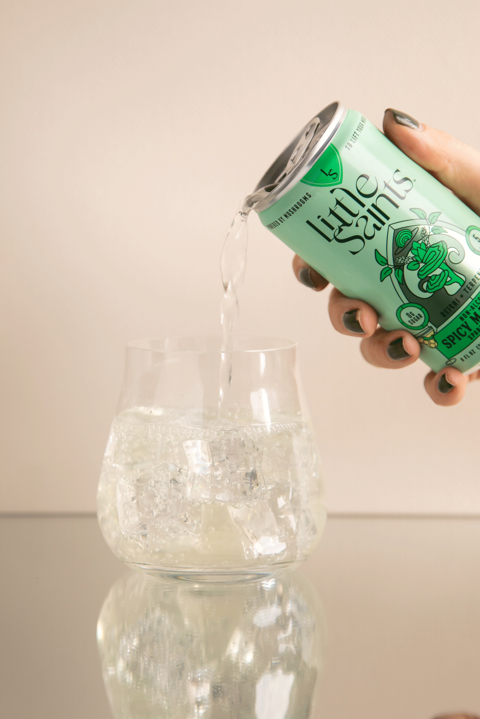 A hand pours a clear drink from a mint-green Little Saints Spicy Margarita Adaptogenic Mocktail can into an ice-filled glass; this non-alcoholic beverage, crafted with reishi mushroom, sits on a reflective surface against a neutral background.