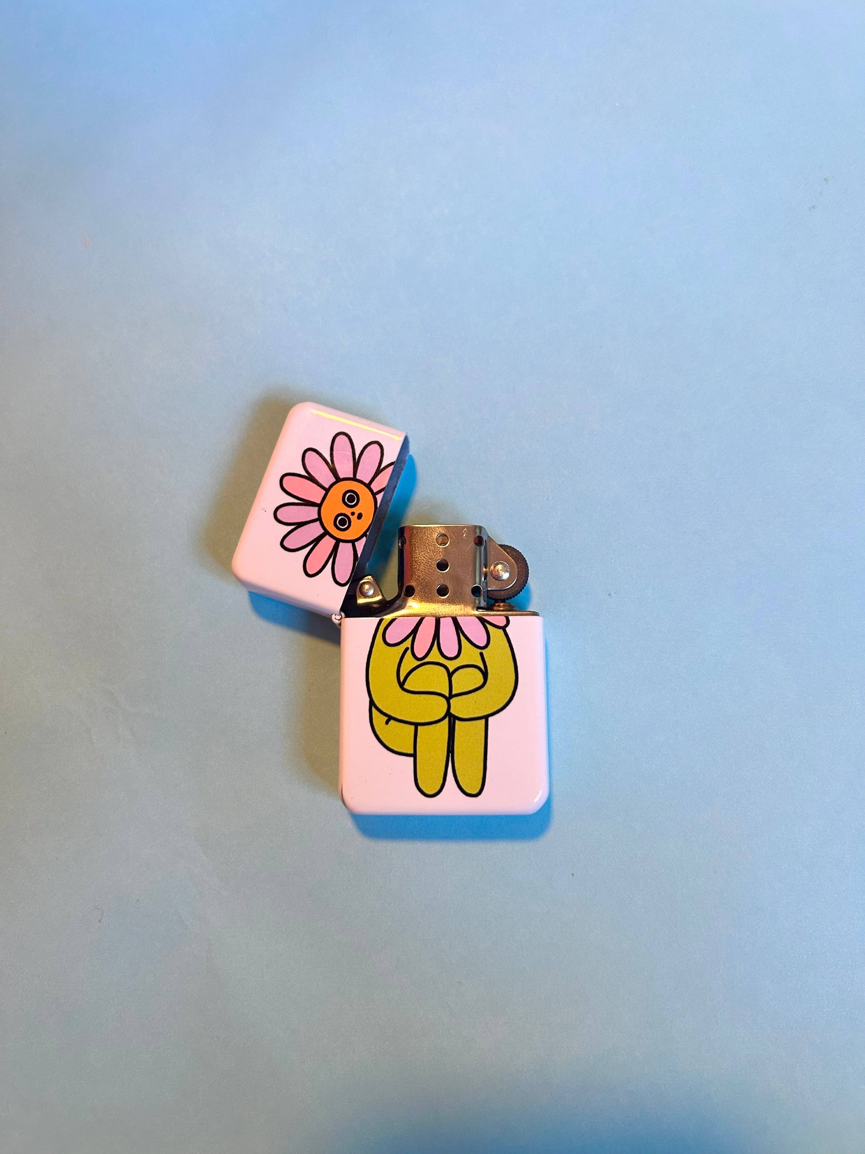 The Peach Fuzzs Flower Boy Lighter: Perfect—a white refillable lighter with a smiling pink flower lid and green hugging cartoon figure—is the ideal gift for stoners or candle lovers, shown on a light blue background.