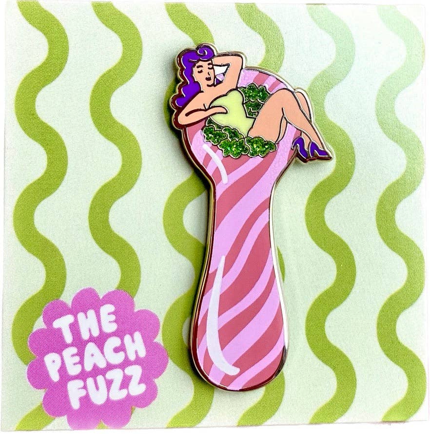 The Peach Fuzzs Total Smoke Show Enamel Pin features a retro pinup girl with purple hair lounging on a pink-striped peach, surrounded by green leaves. A wavy green background highlights the brand name in a purple flower shape.