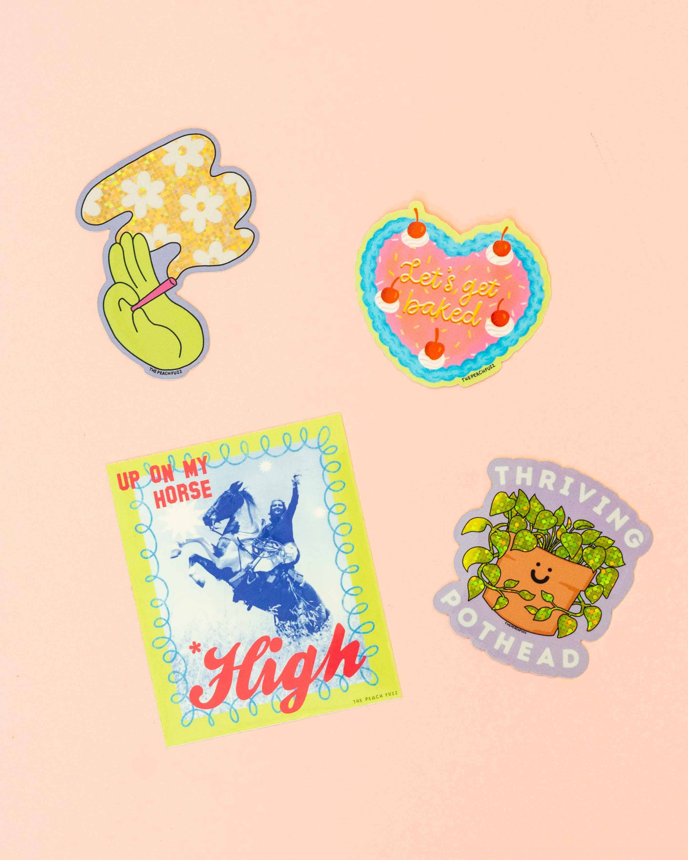 Four colorful waterproof The Peach Fuzz stickers on light pink: a daisy peace sign, Let’s Get Baked Cake Sticker, a cowboy with “High,” and a smiling taco with “Thriving Pothead.” Perfect for laptops!.