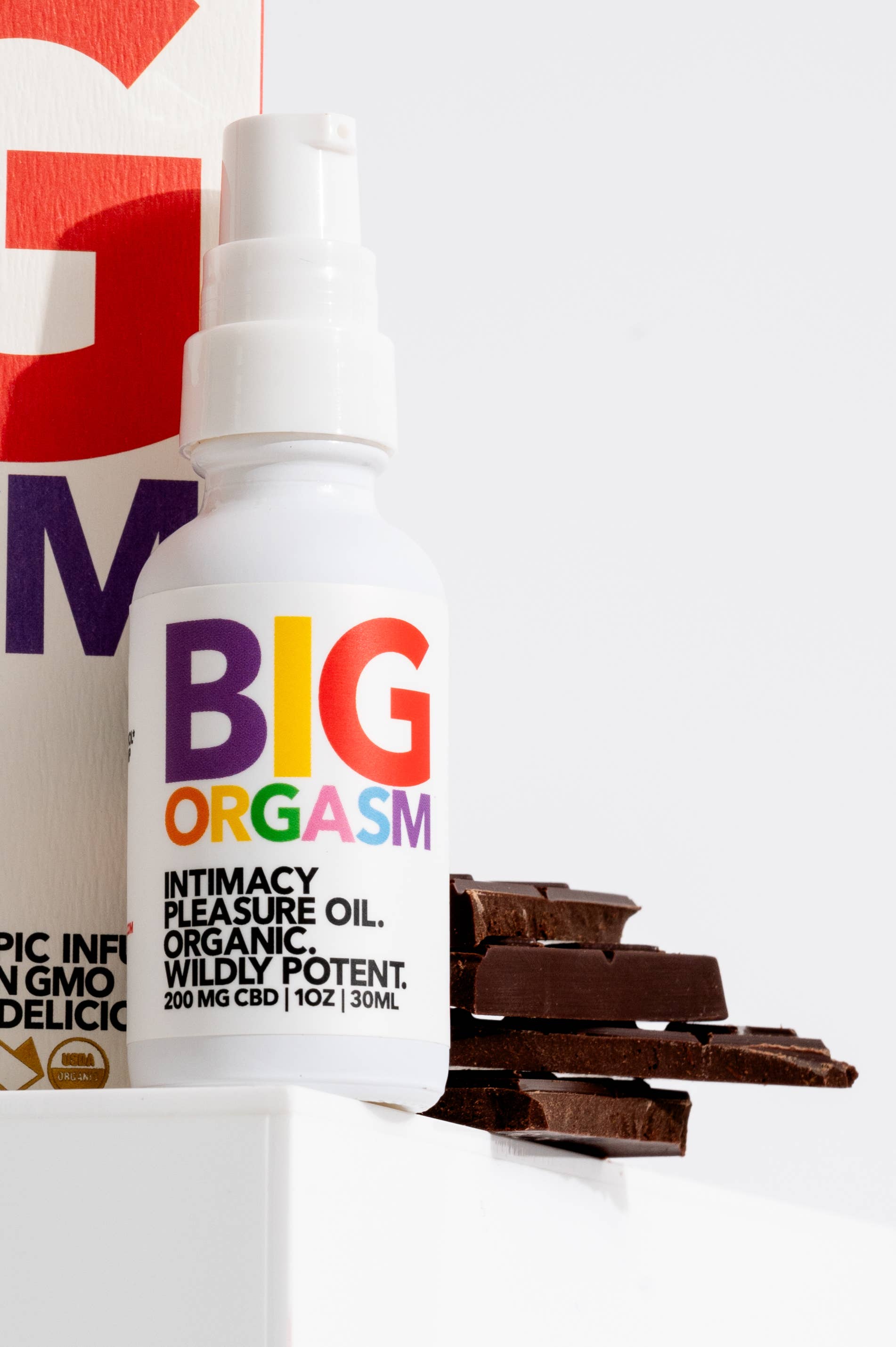 A white bottle labeled Big Orgasm Oil by Eat Gold Organics stands upright next to chunks of dark chocolate and its box. This organic arousal oil features damiana leaf, all presented on a crisp white background.