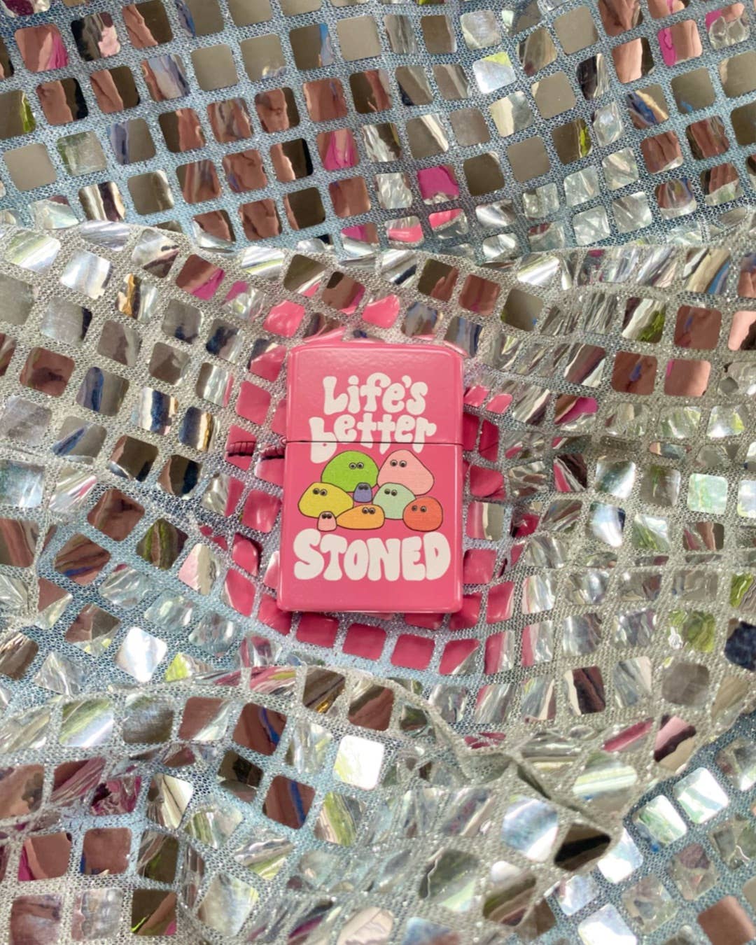 The Peach Fuzz Lifes Better Stoned Lighter, an eco-friendly pink lighter with cartoon faces and the text Lifes Better Stoned, sits on a shiny, reflective silver mosaic surface.