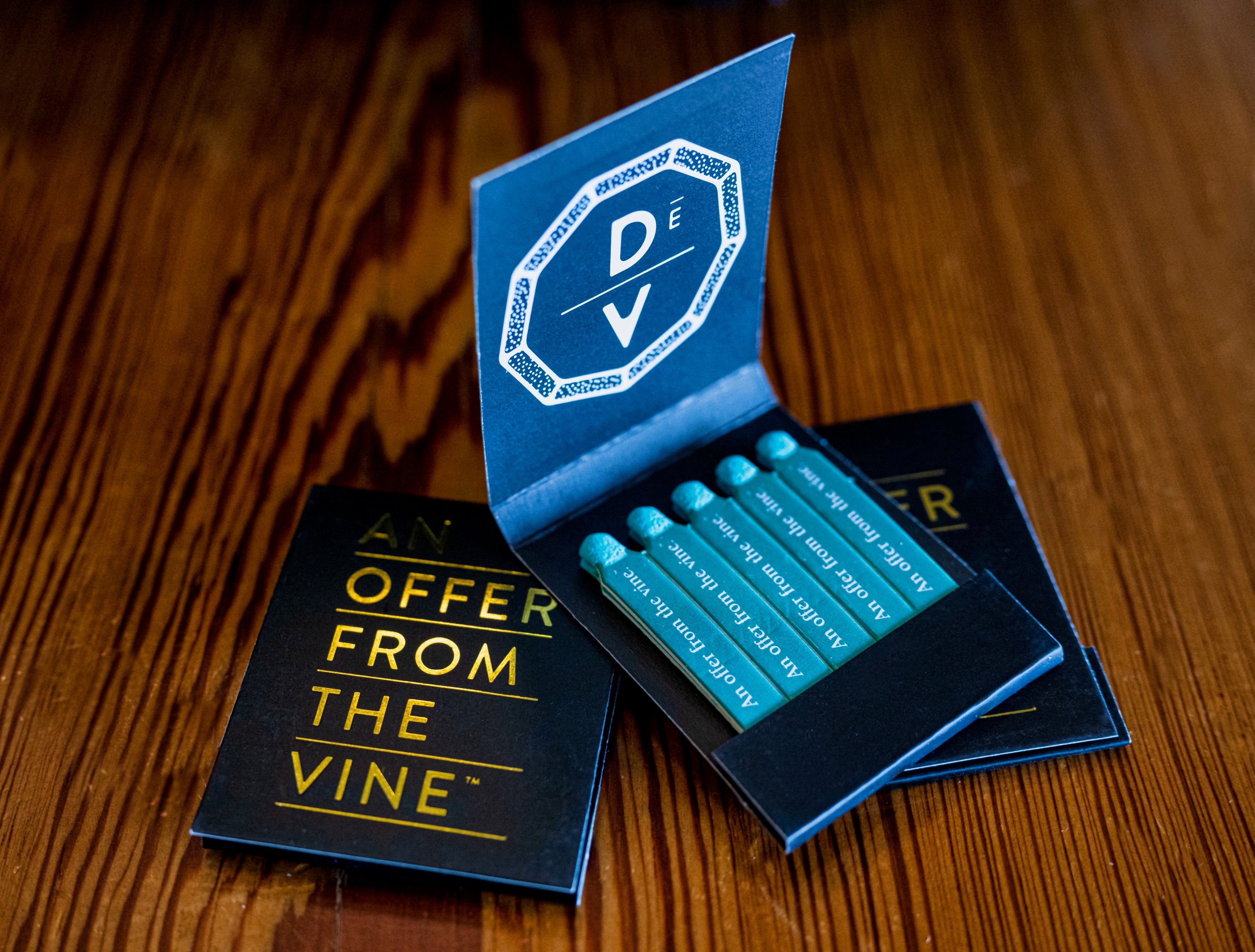 A close-up of stylish deVINE Matches from DeVINE Wellness, featuring teal-tipped matches and an open cover with a white geometric DÊ V logo—perfect for enhancing your herb or smoking experience on a wooden surface.