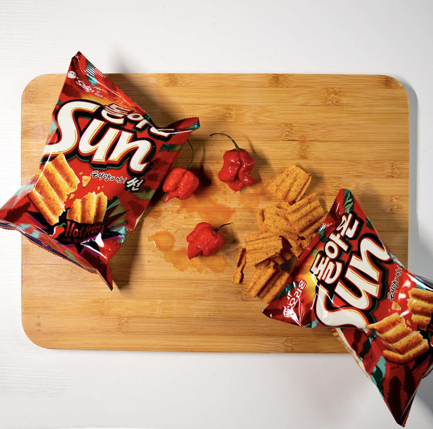 Two bags of Exotic Snacks Sunchips Crazy Hot! Flavor, several red chili peppers, and spicy chips imported from South Korea are arranged on a wooden cutting board atop a white surface.