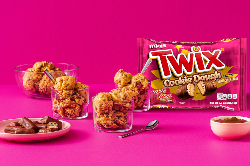 Bowls and glasses of cookie dough ice cream rest on a pink surface, surrounded by Exotic Snacks Twix - Cookie Dough Minis and chopped milk chocolate Twix bars, all set against a vibrant pink background.