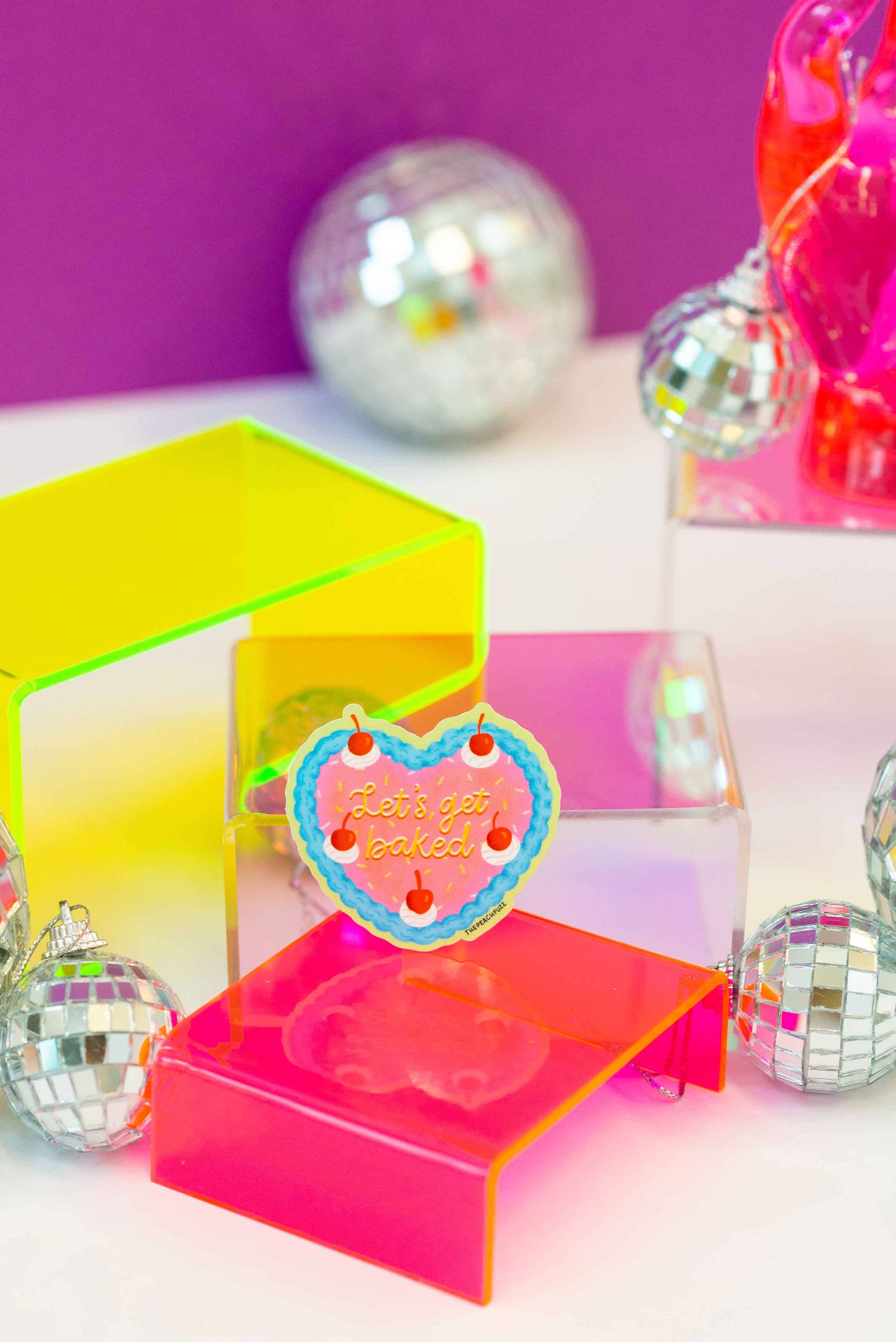 The Peach Fuzzs Lets Get Baked Cake Sticker features a heart-shaped cookie with playful text, shown among neon acrylic stands and disco balls on a purple background—ideal as a waterproof baking sticker for your laptop.