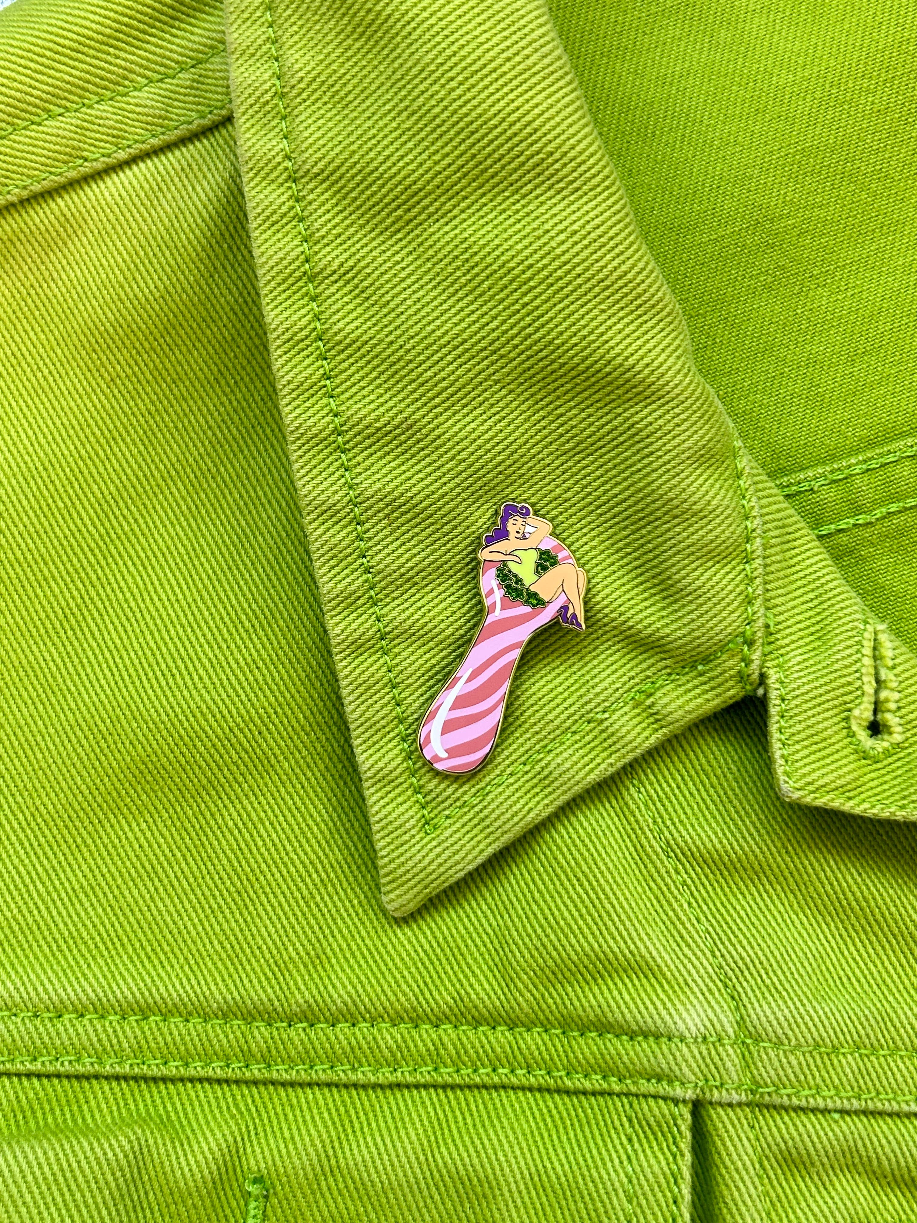 The Peach Fuzz’s Total Smoke Show Enamel Pin, featuring a cartoon woman in a green outfit and pink-striped stockings, is attached to the collar of a bright green denim jacket.