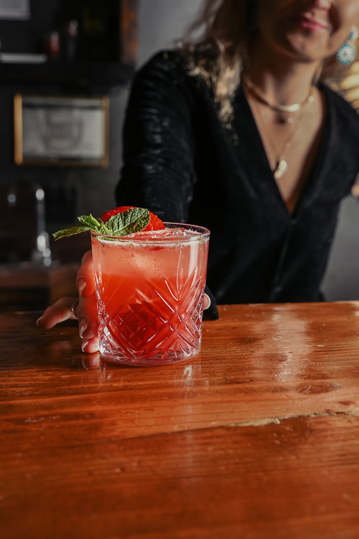 Woman in black serving a red drink garnished with mint called Awake Zero Proof Elixir on a wooden bar counter