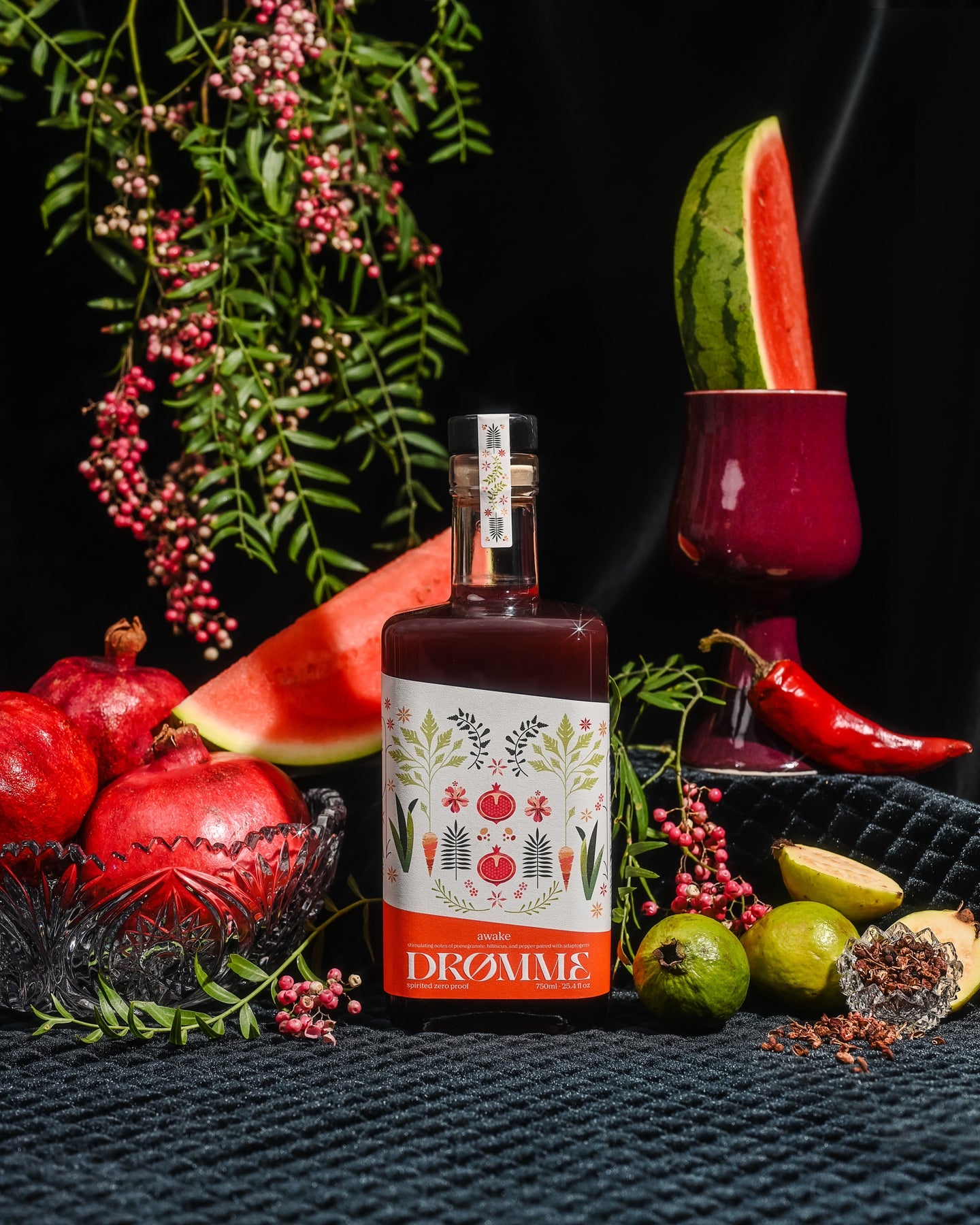 Bottle of Awake Zero Proof Elixir surrounded by pomegranate, watermelon, lime, chili, and peppercorns on a dark surface.