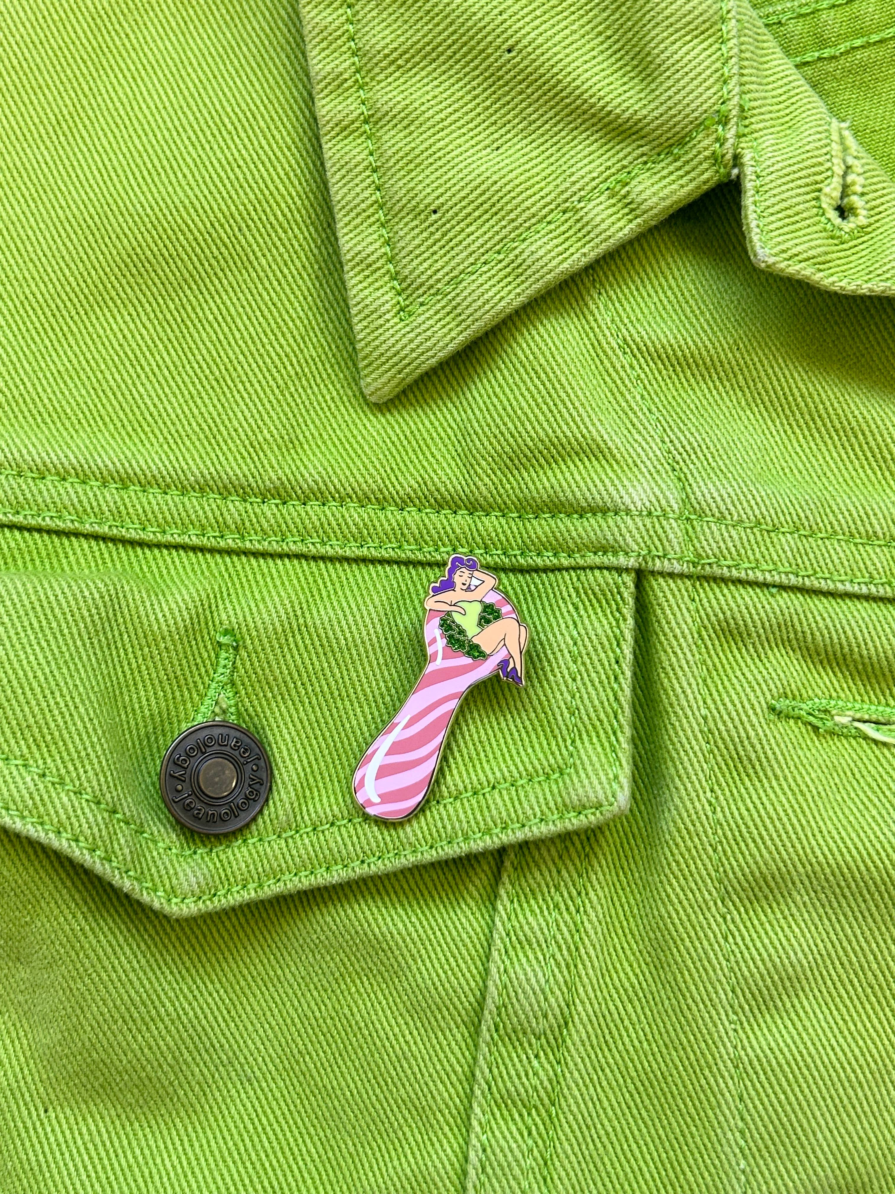 The Peach Fuzzs Total Smoke Show Enamel Pin—a colorful glitter sock with pink stripes and a cartoon frog—adds a playful touch to the pocket of a bright green denim jacket.