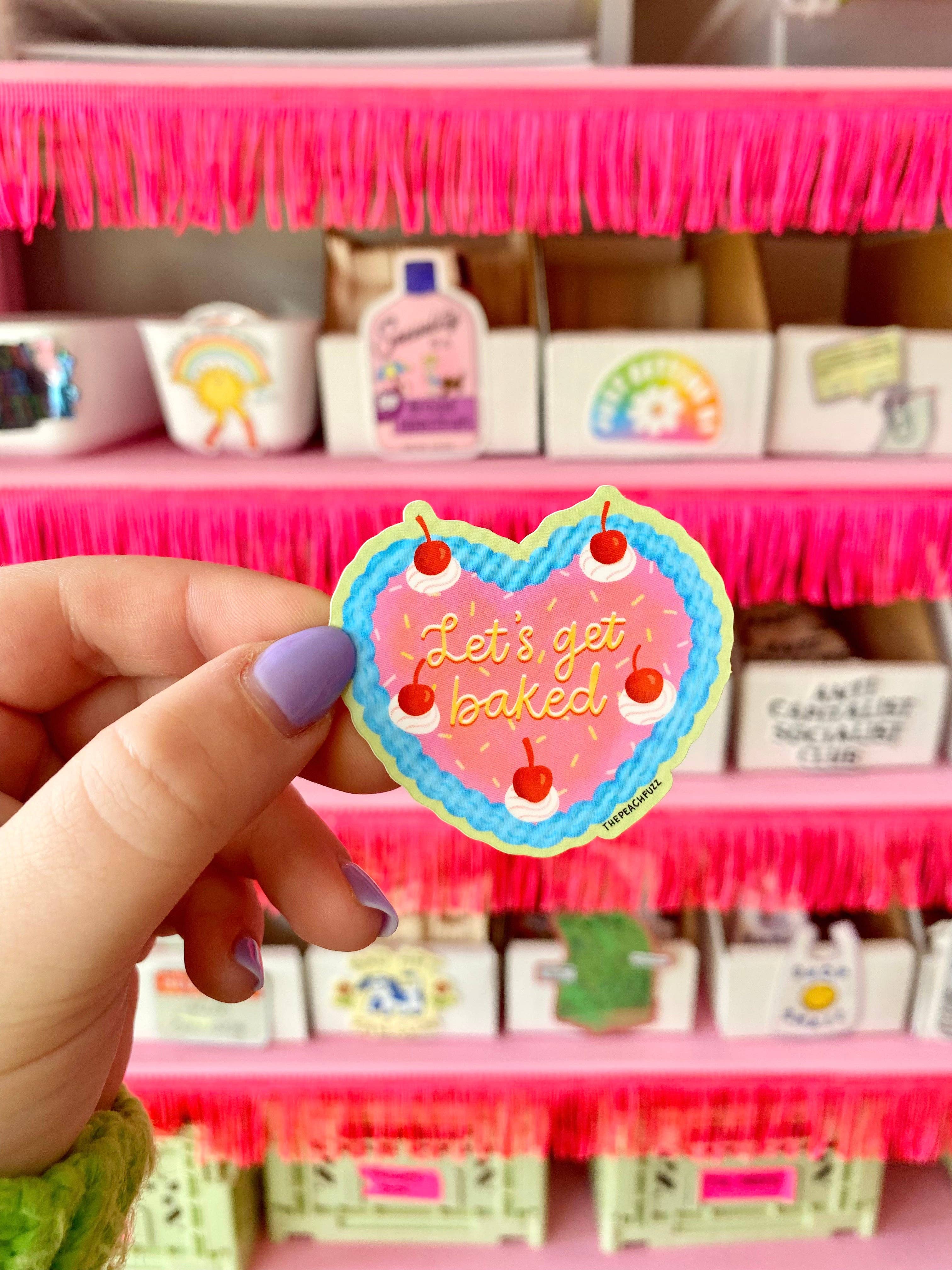 A hand with lavender nail polish holds The Peach Fuzz Lets Get Baked Cake Sticker. The heart-shaped sticker features cherries and the phrase Let’s Get Baked, with pink shelves and colorful bins in the background.